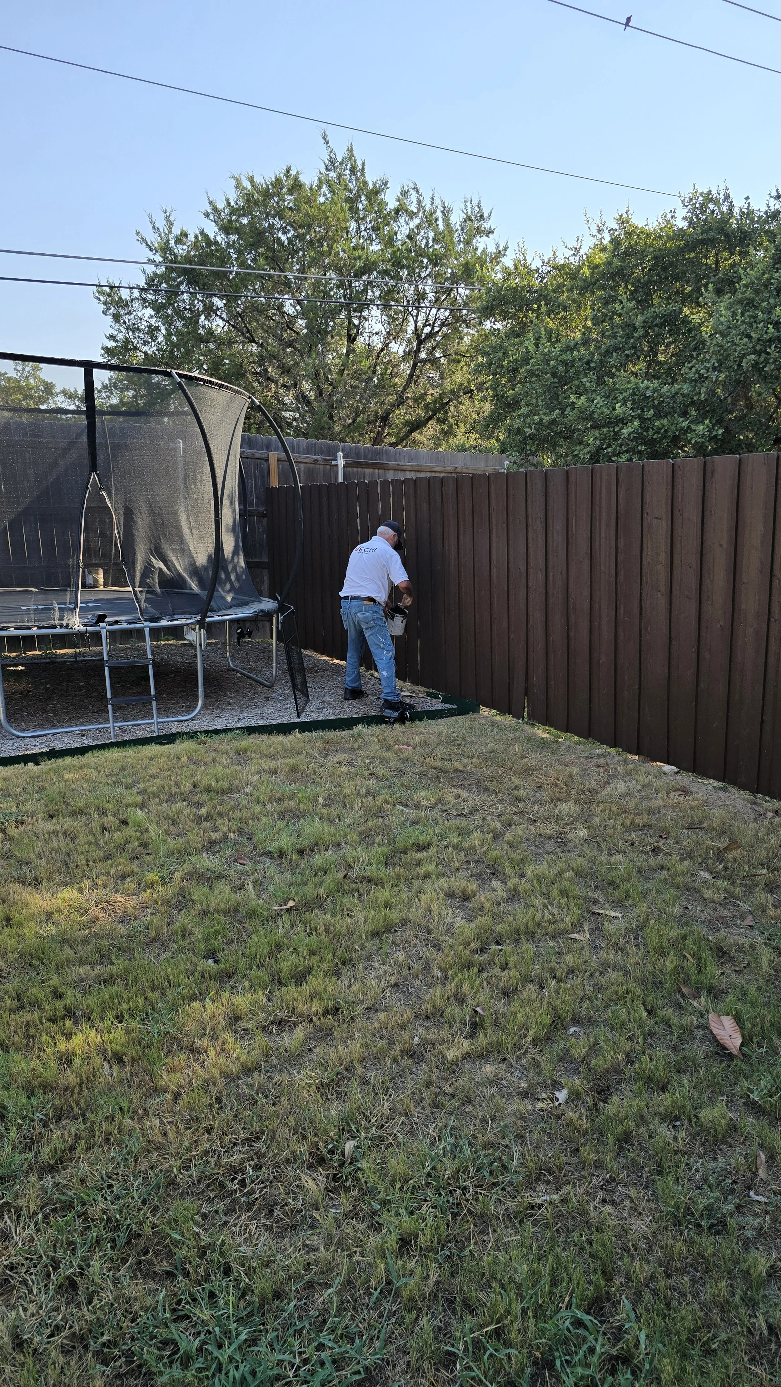 A man wearing a white shirt and blue jeans is installing a dark brown wooden fence in a backyard. There is a trampoline with a safety net on the left side and some trees in the background.