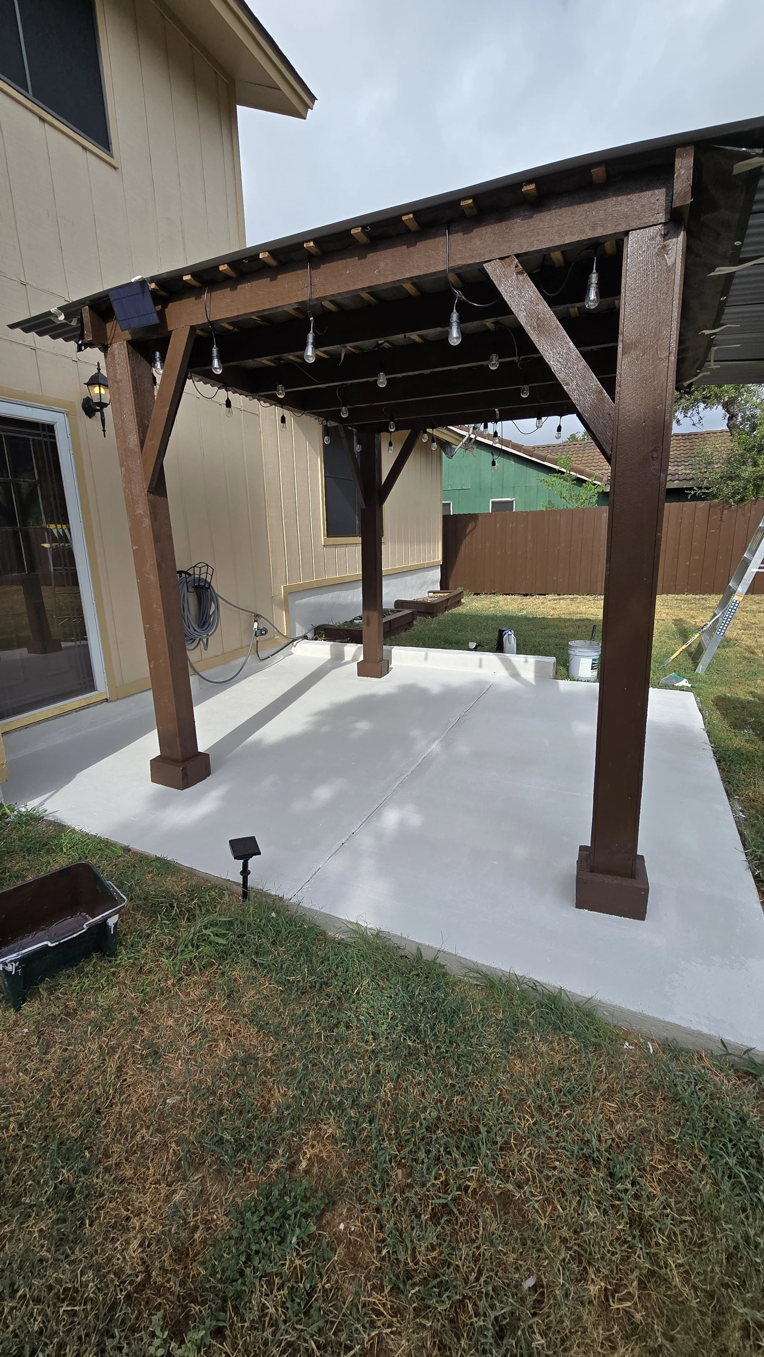A newly constructed wooden patio cover with string lights sits over a concrete patio in a backyard, with a house, grass, and a green shed in the background.