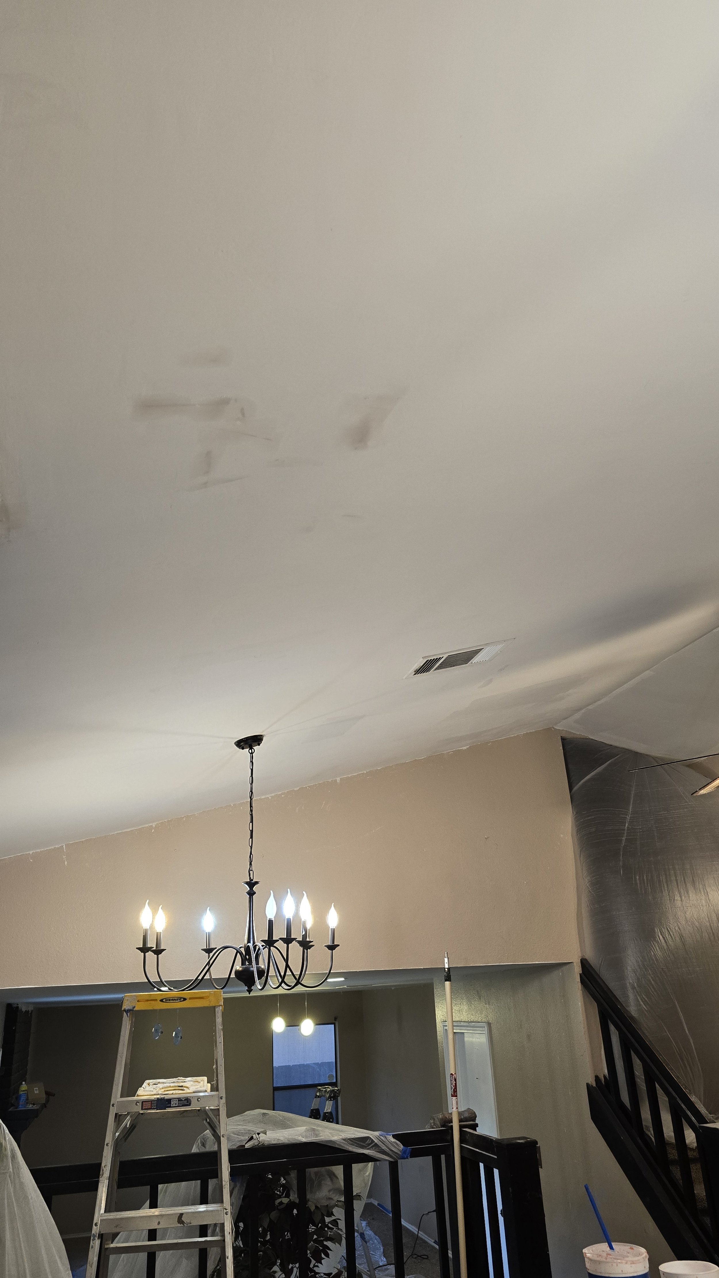 A room under renovation with a chandelier, a ladder, and construction tools. The ceiling has patches of drywall repair.