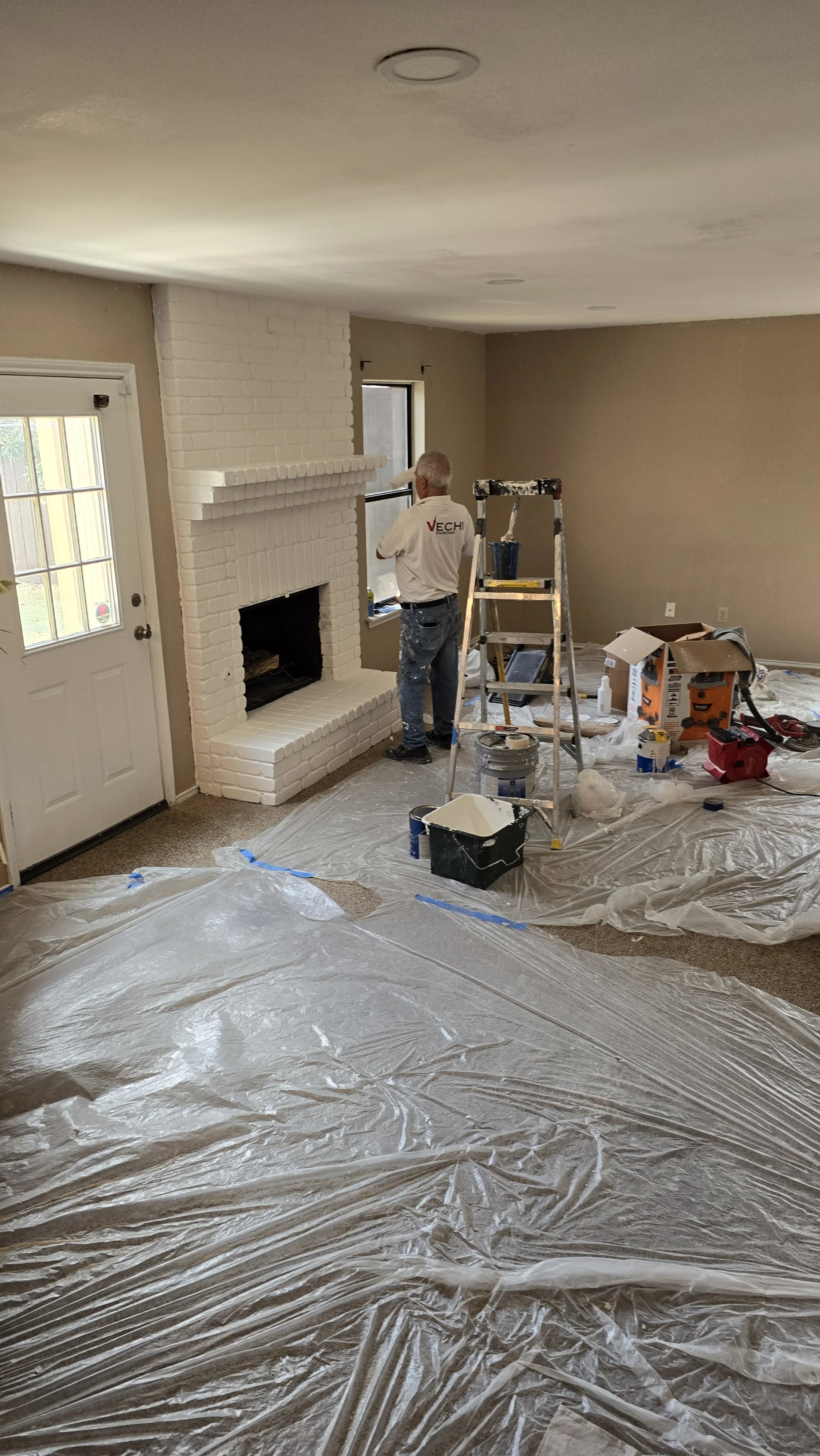 A person painting a white fireplace in a living room, with various painting supplies and tools scattered on the floor covered with plastic.