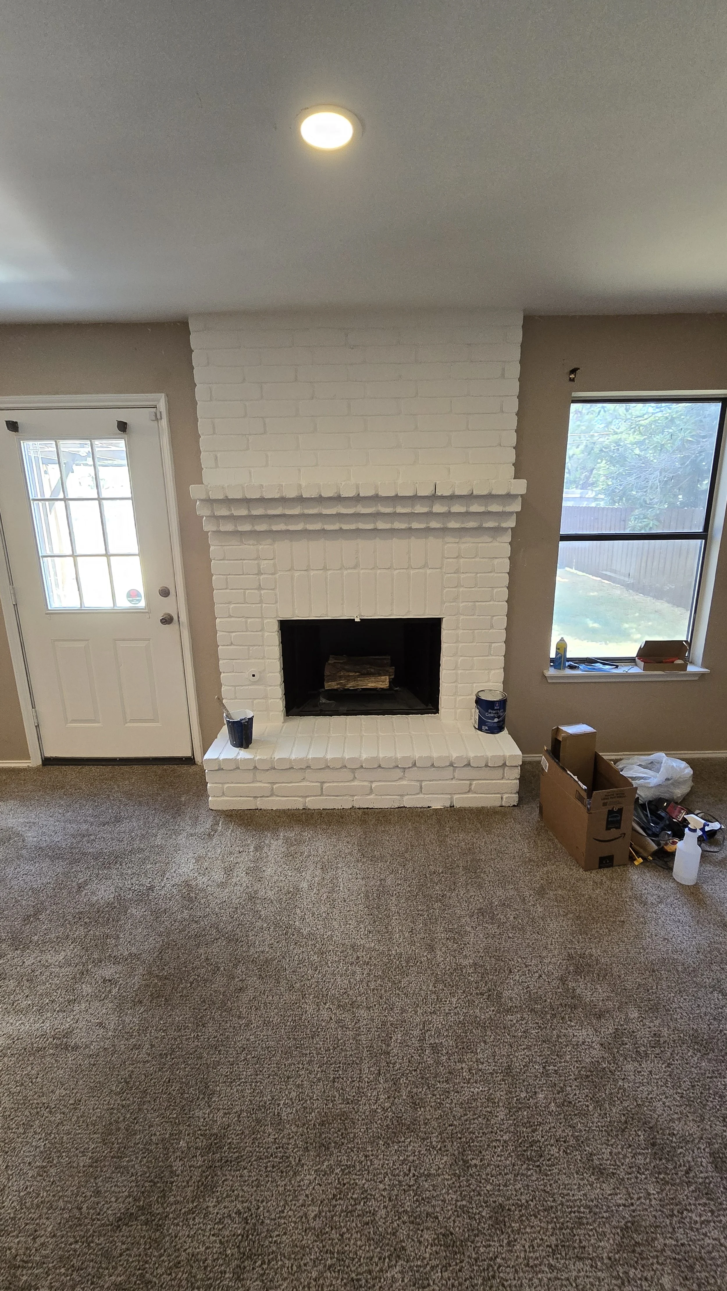 Living room with white painted brick fireplace, open hearth with logs, beige carpeted floor, window to the right, door to the left, and boxes and tools on the floor nearby.