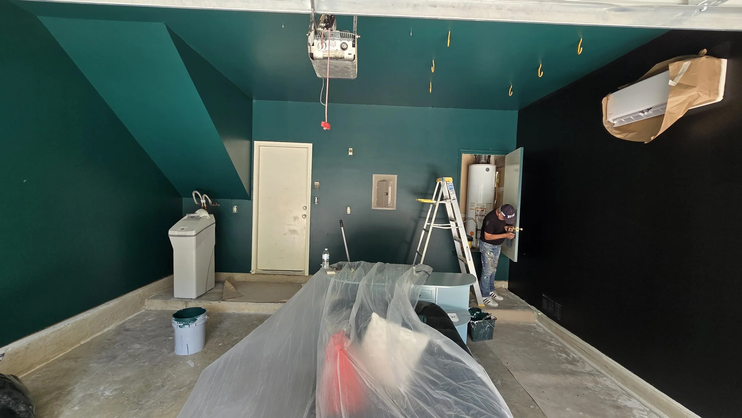 Interior room under renovation with a person working near a ladder, paint cans, plastic cover, water heater, and air conditioning unit.