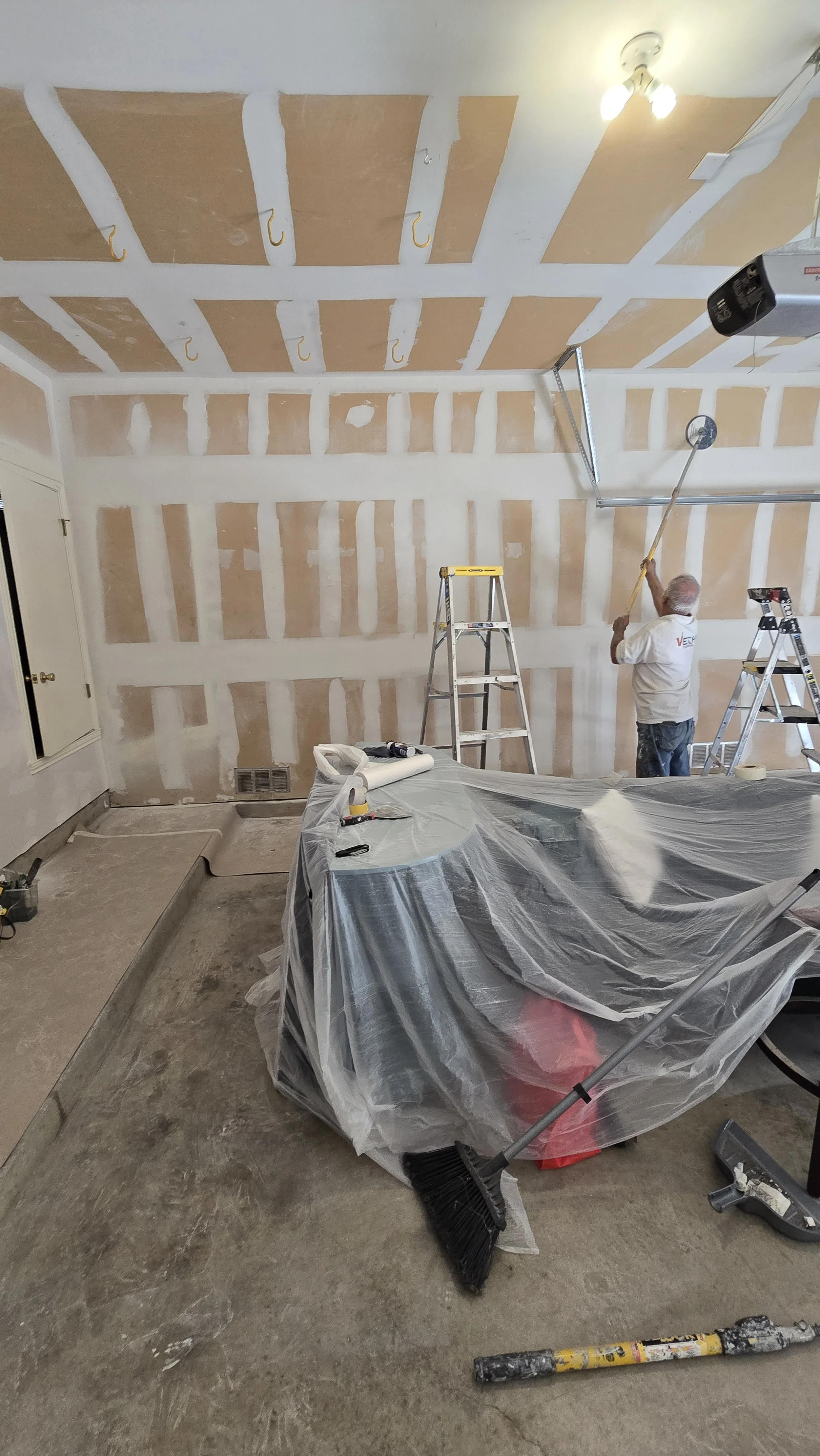 A person working on a drywall ceiling with multiple hooks, using a long pole, in a room under construction, with ladders, tools, and a covered sofa.