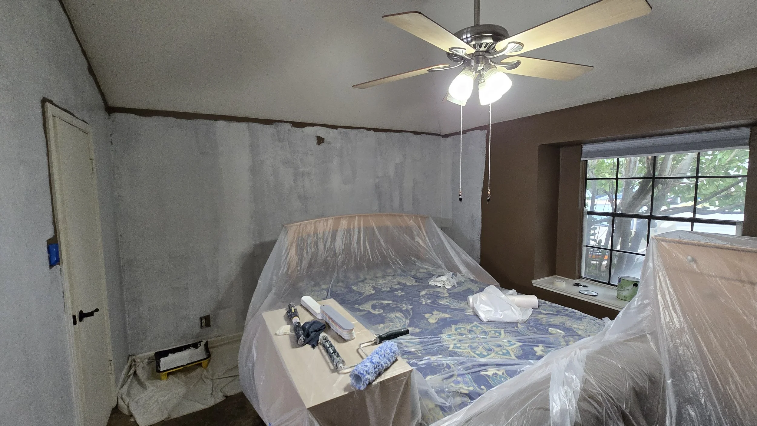 Room undergoing painting renovation with plastic covering furniture, painting tools on a table, a ceiling fan, and a window with trees outside.
