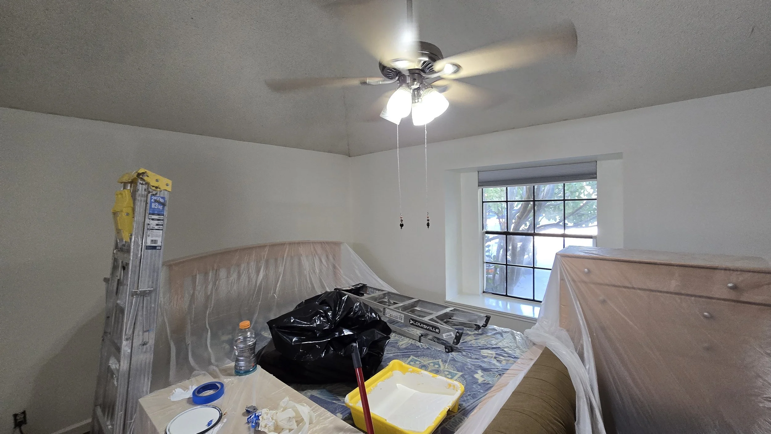 Room under renovation with ladder, plastic coverings, and painting supplies