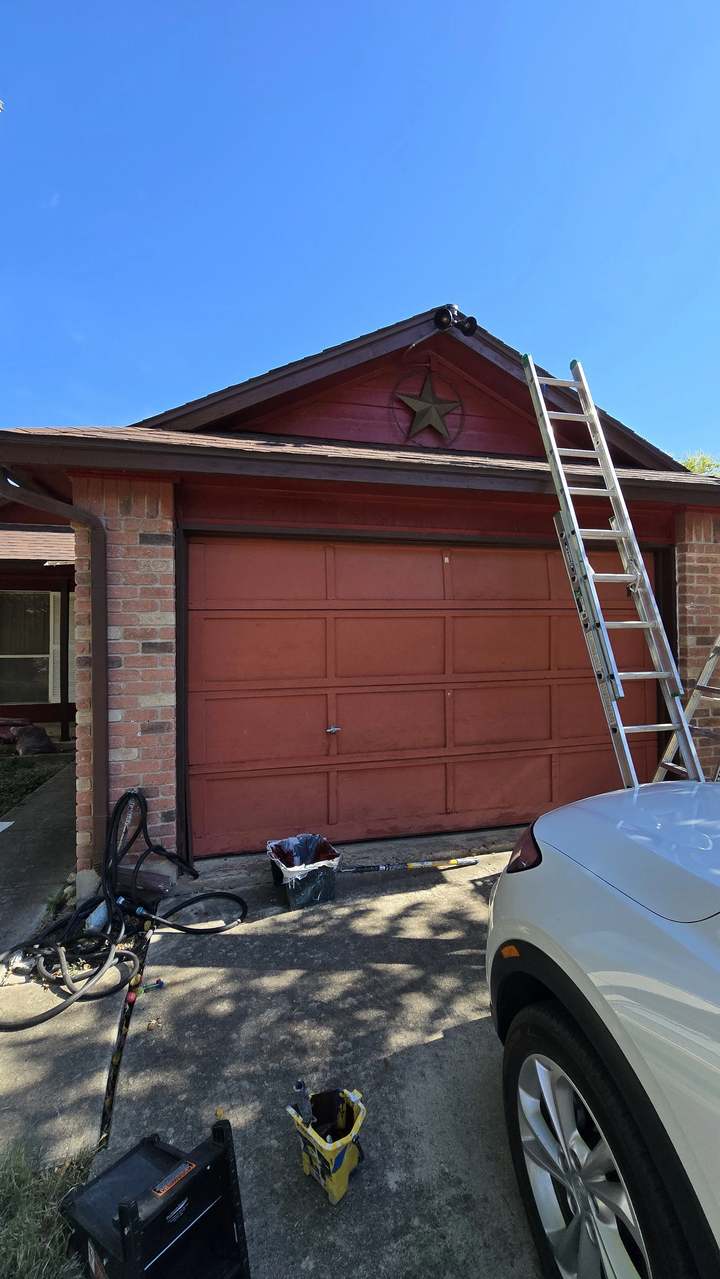Red garage door with a star emblem on the gable above, ladder leaning against the house, car parked in driveway, and cleaning tools and supplies on the ground.