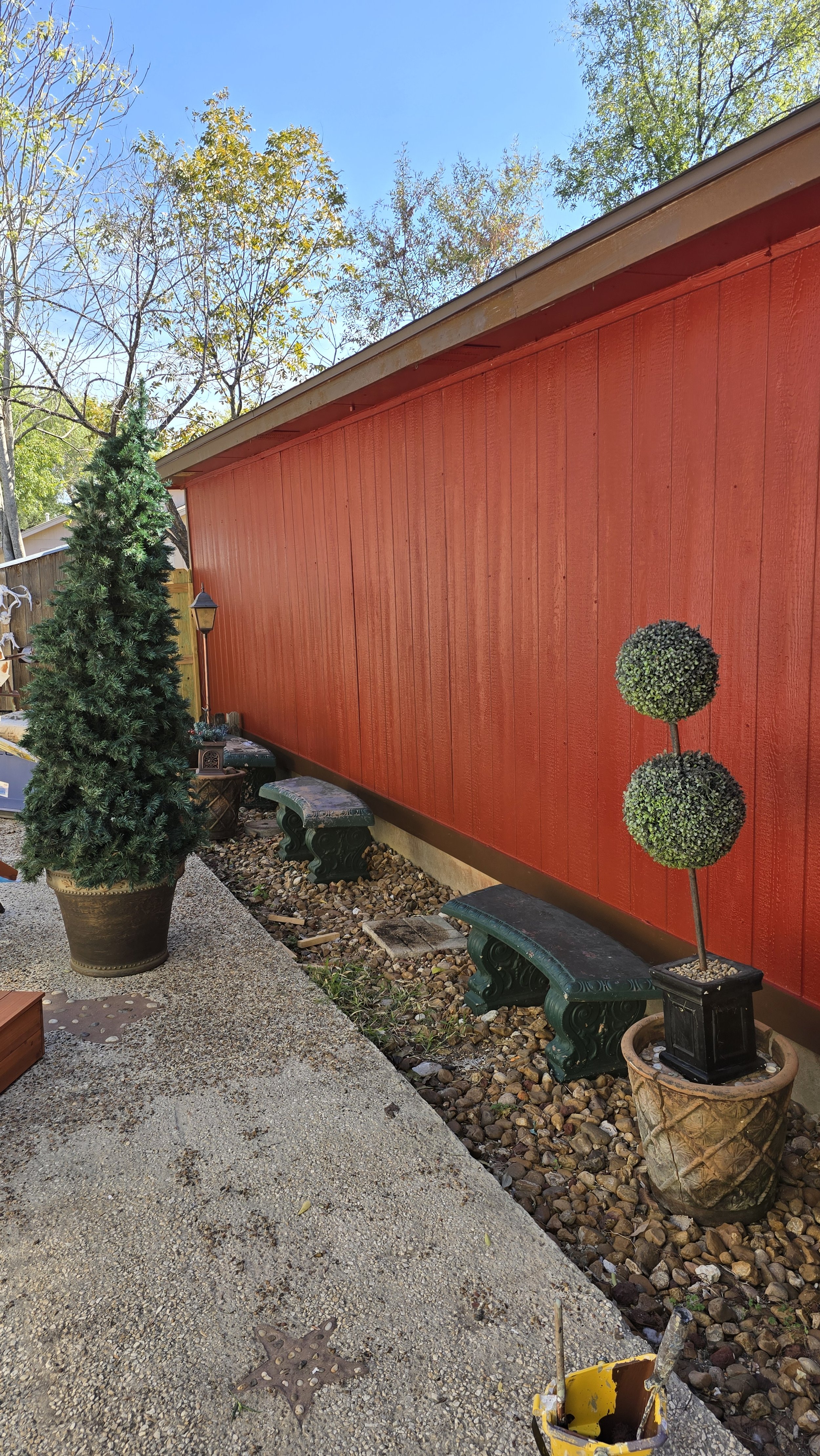 A backyard scene featuring a red wooden fence, a large potted evergreen tree, decorative topiary, and outdoor benches with stone and gravel ground, under a clear blue sky.