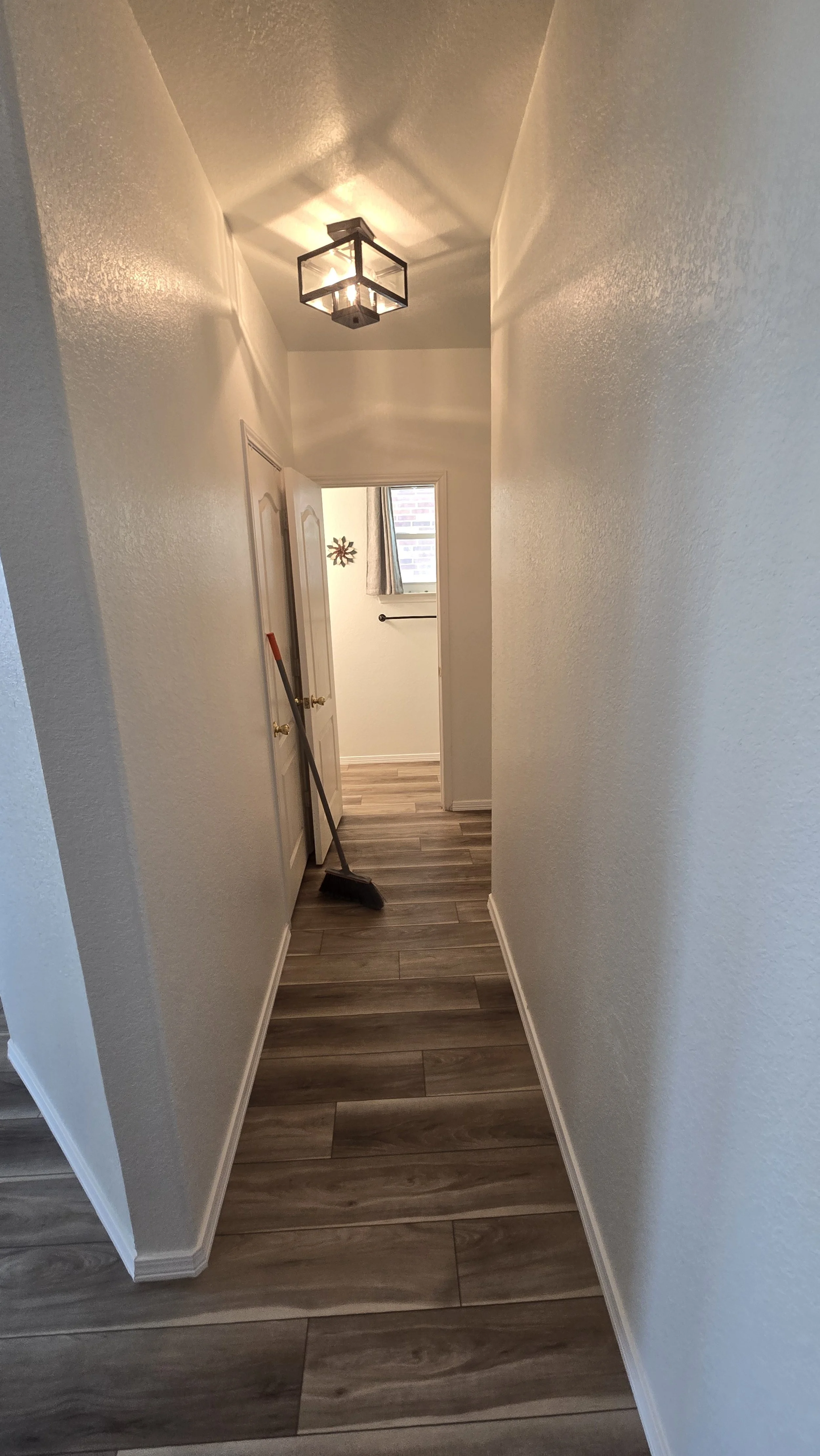 A narrow hallway with white walls, a wood-look floor, and a ceiling light fixture. A broom is leaning against a door on the left side, and a small window with curtains is at the end of the hallway.
