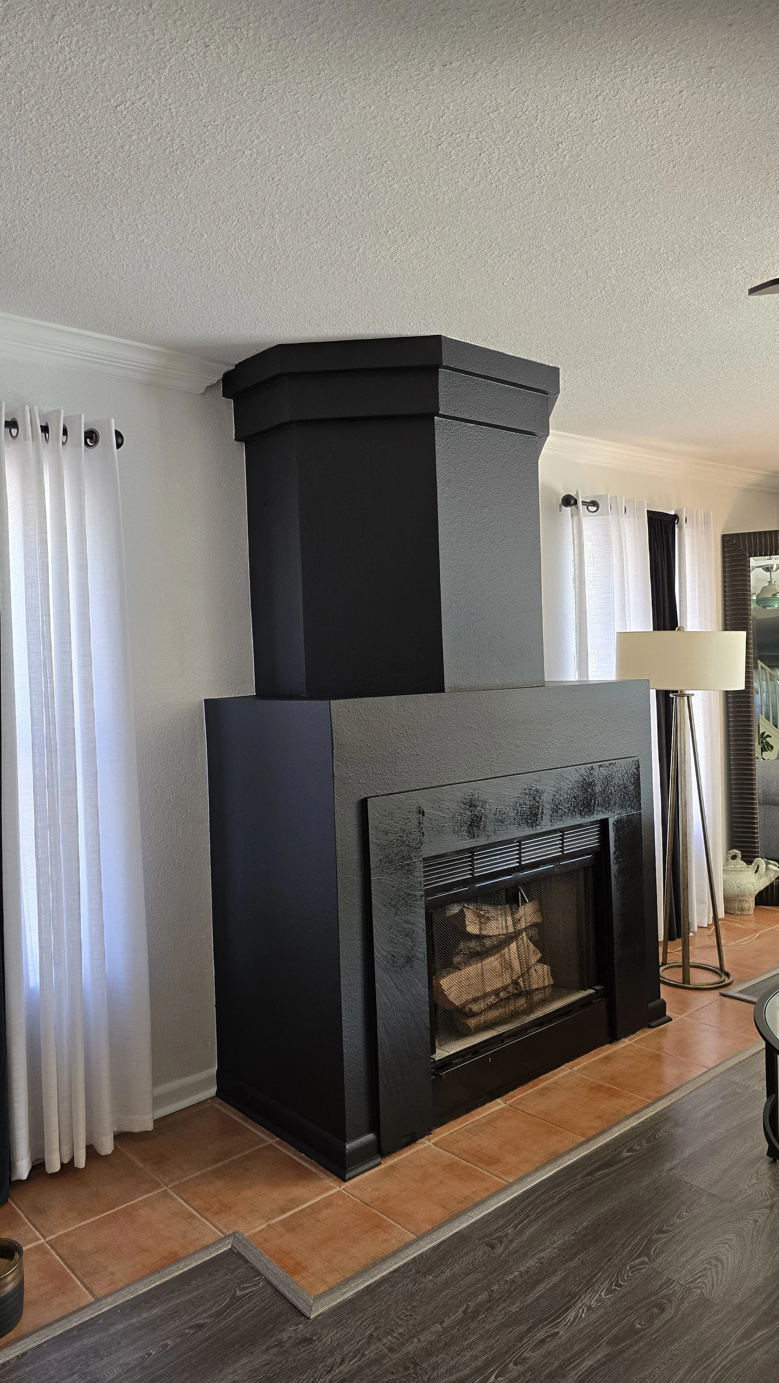 Black painted fireplace with a protruding chimney in a living room, white curtains on either side, a standing lamp, and part of a dark wood table in the foreground.