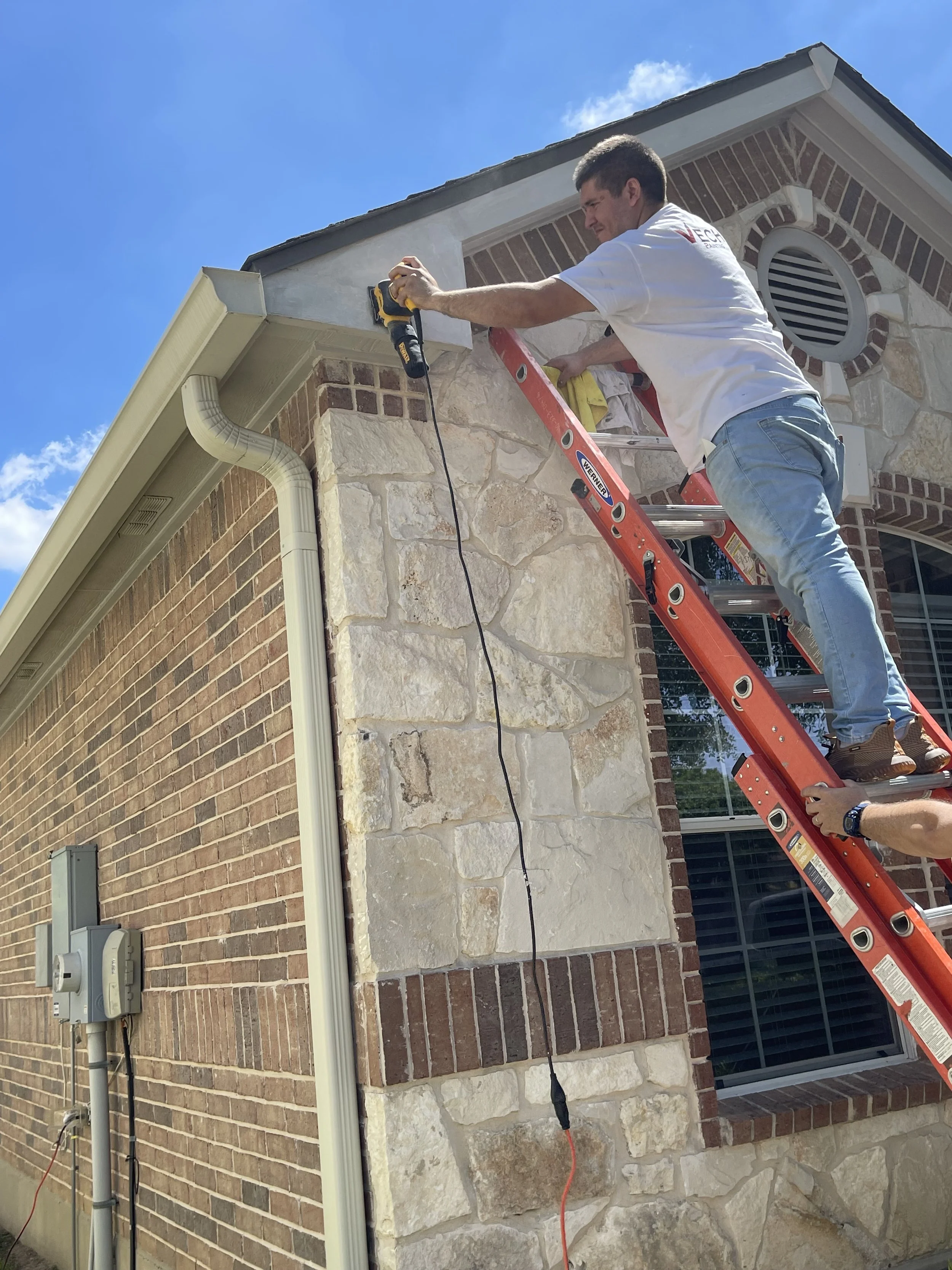 A man on a ladder cleaning the eaves of a house with a power washer under a blue sky.