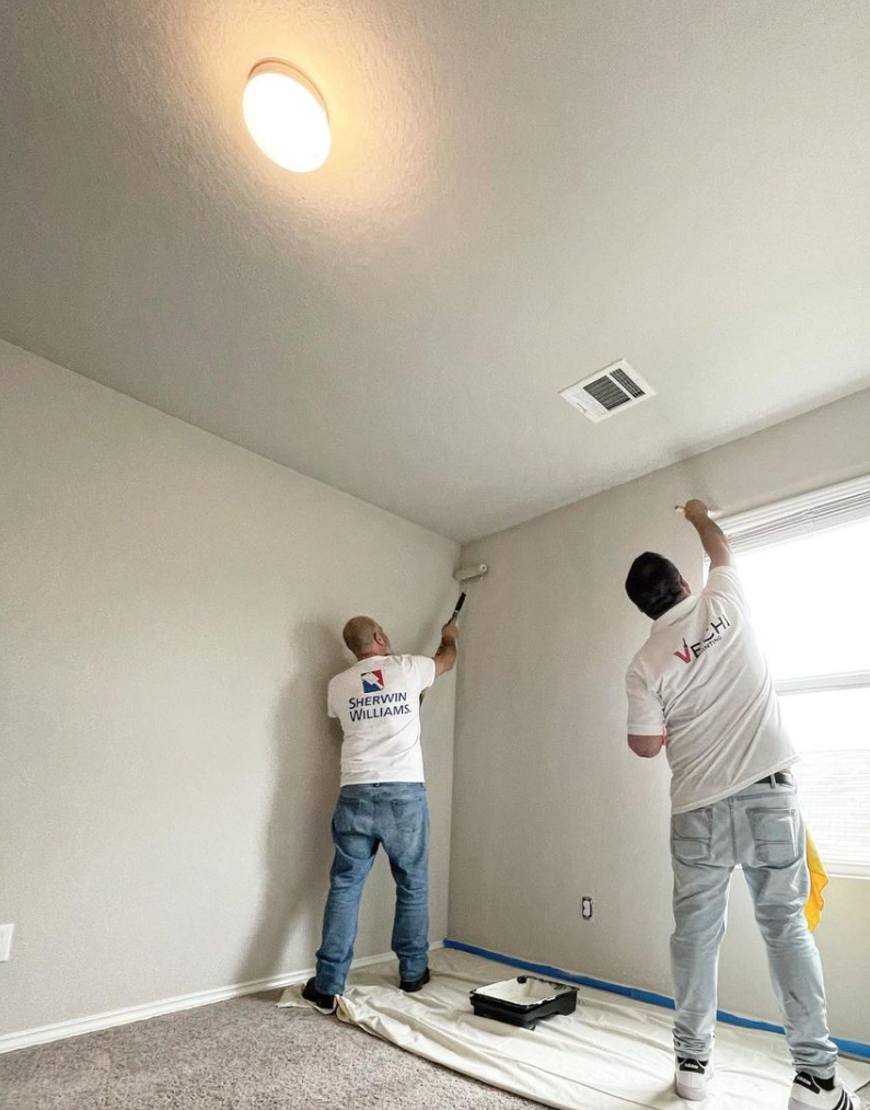 Two workers painting a wall in a room, one using a roller on an extension pole and the other using a brush or roller near the ceiling, with a drop cloth on the floor.