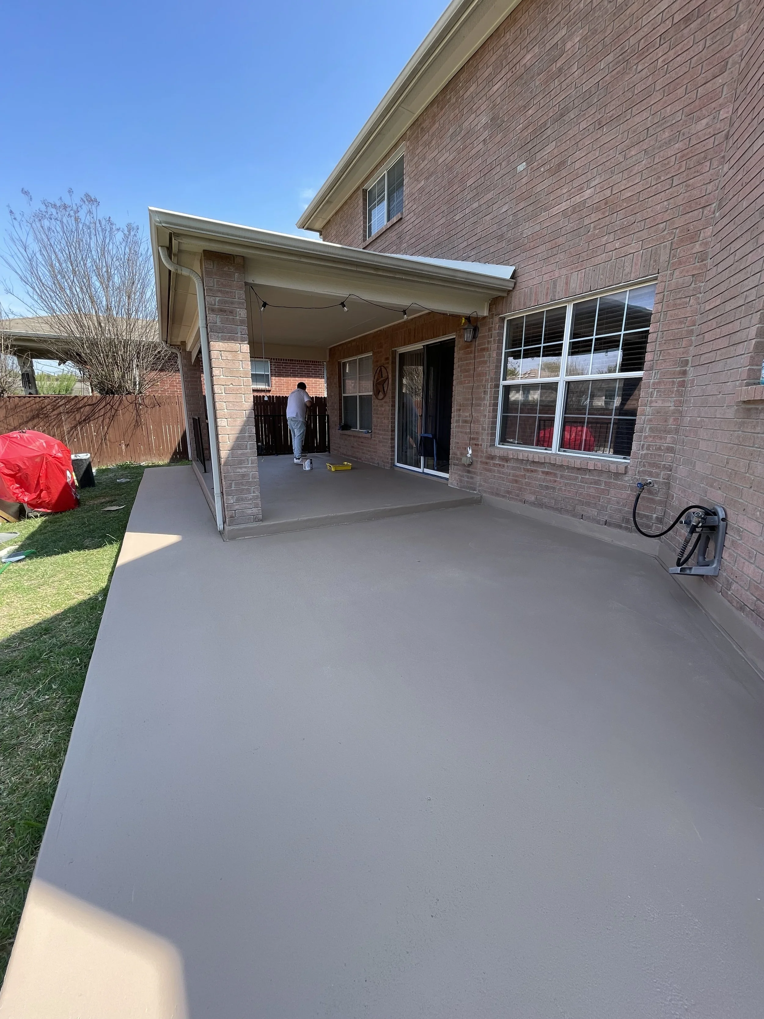 Backyard patio with concrete flooring adjacent to a brick house, person painting the wall, with a fence and tree in the background.