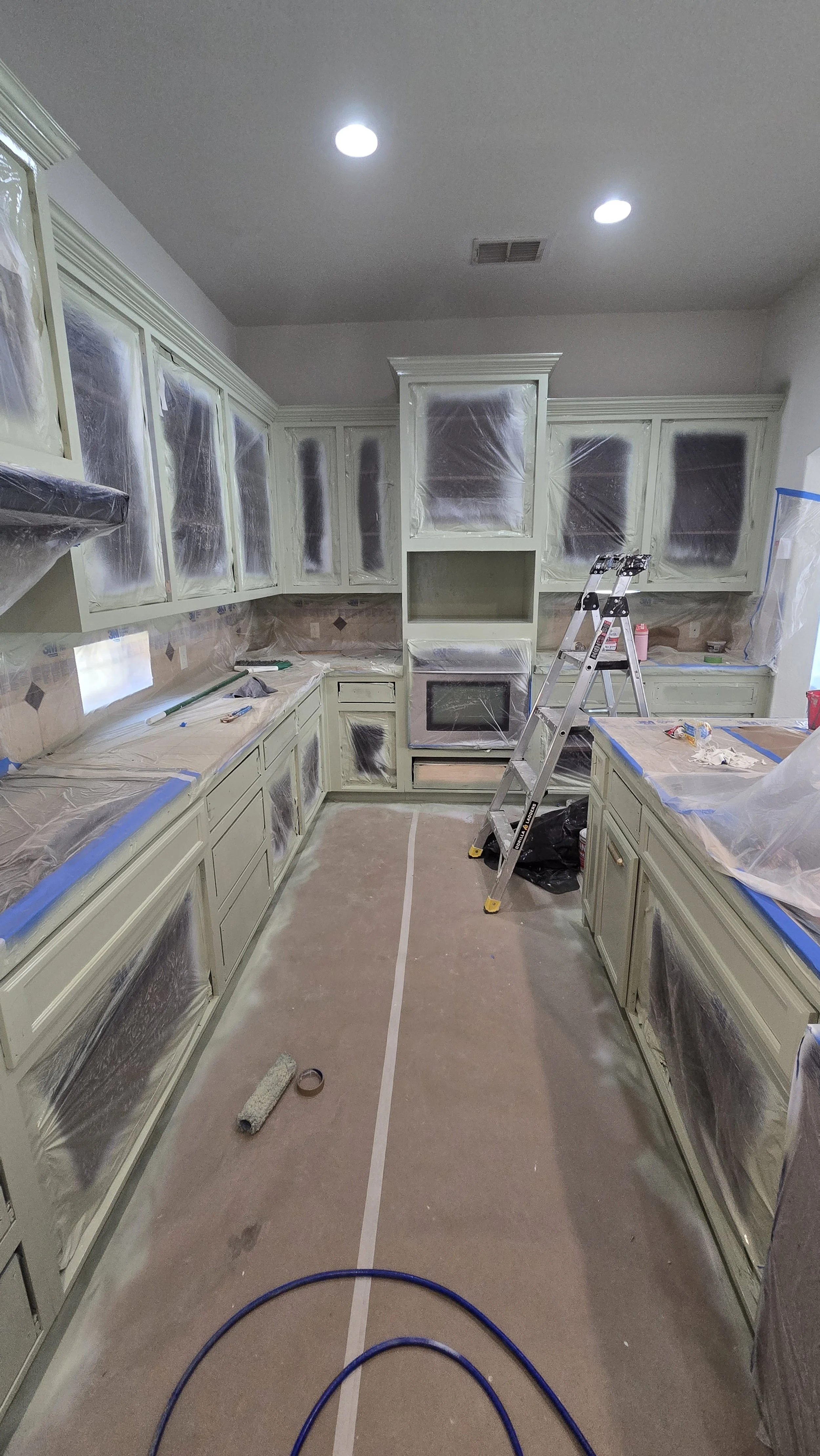 Kitchen undergoing renovation, cabinets and appliances covered with plastic, ladder in the center, construction materials on the floor.