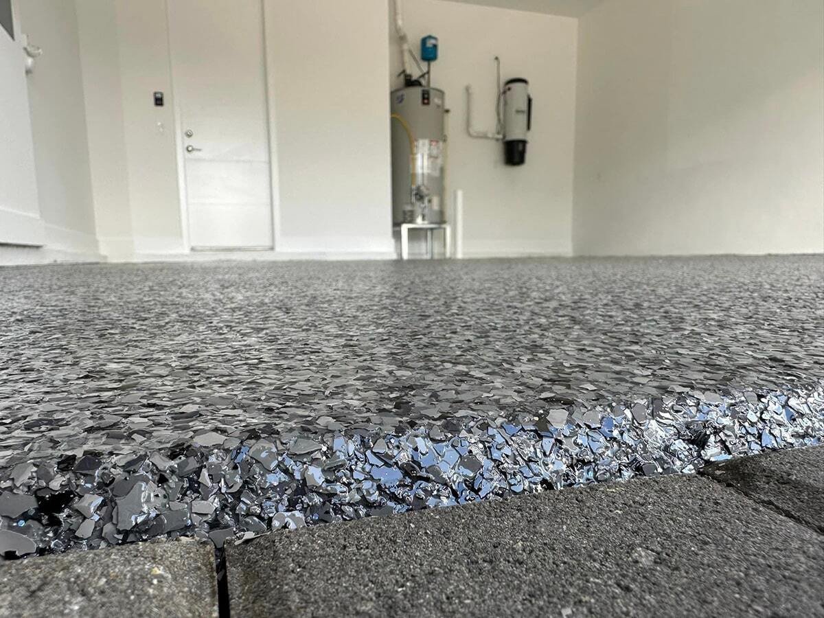 Close-up view of a newly installed epoxy garage floor with a textured finish, showing the transition from bare concrete to coated surface in a garage with white walls and equipment in the background.