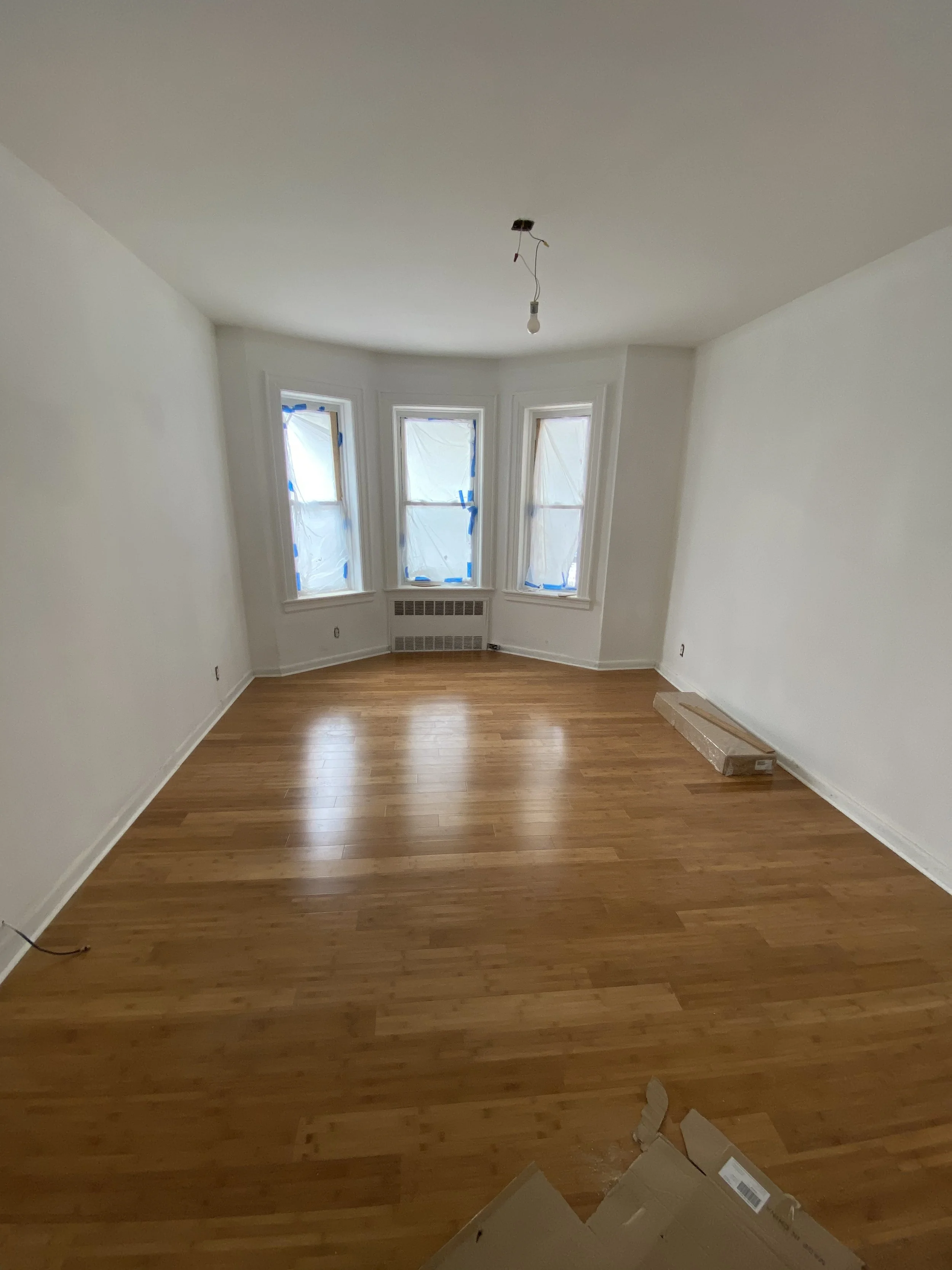 Empty living room with hardwood floors, three windows covered with protective plastic, and a bare light bulb hanging from the ceiling.