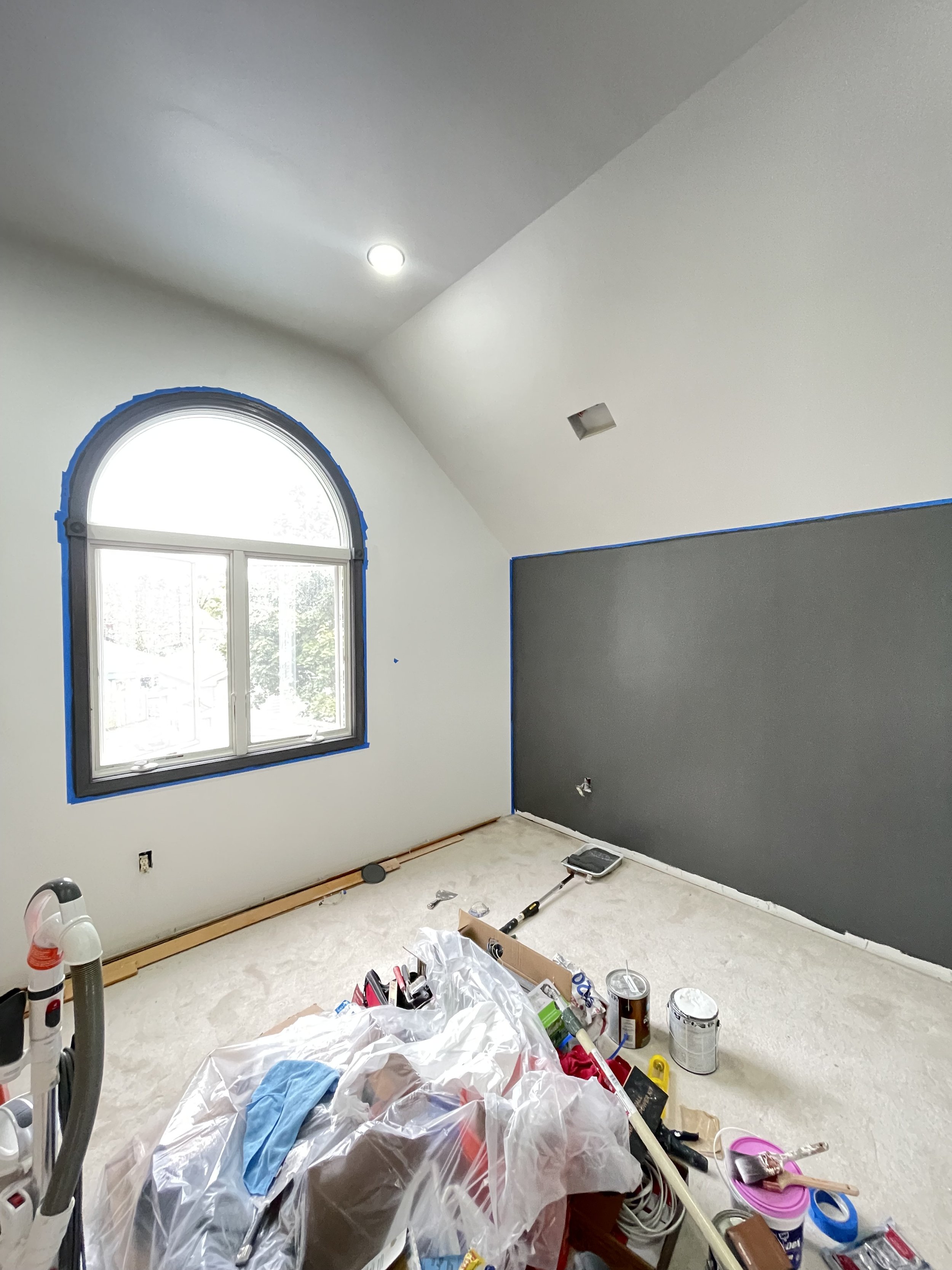 Room under renovation with painting supplies on the floor, an unfinished wall with gray paint, a large arched window, and construction tools.