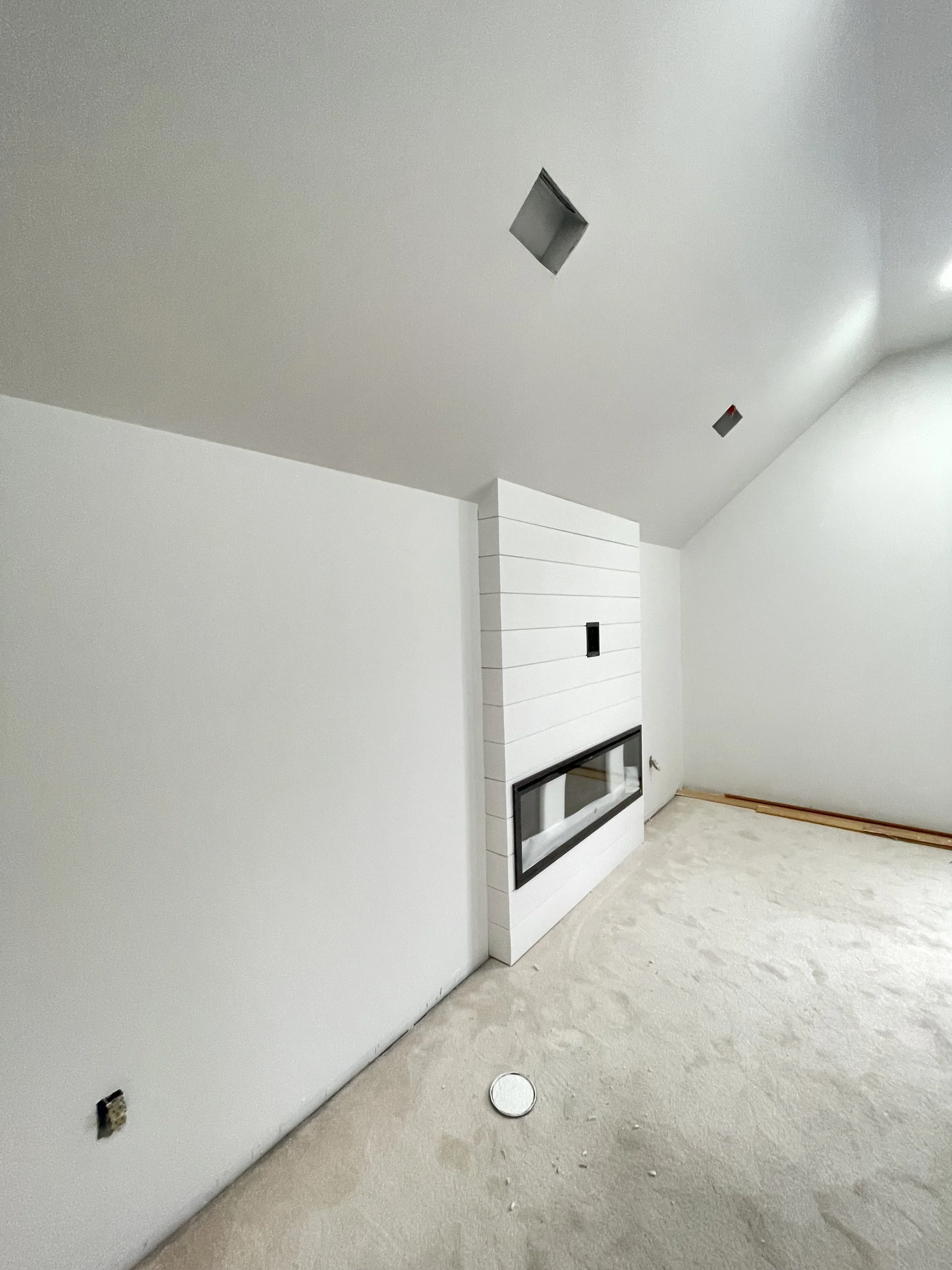 Empty unfinished room with a white wall, ceiling with cutouts for lighting, a white-shiplap fireplace, and a circular access panel on the floor.