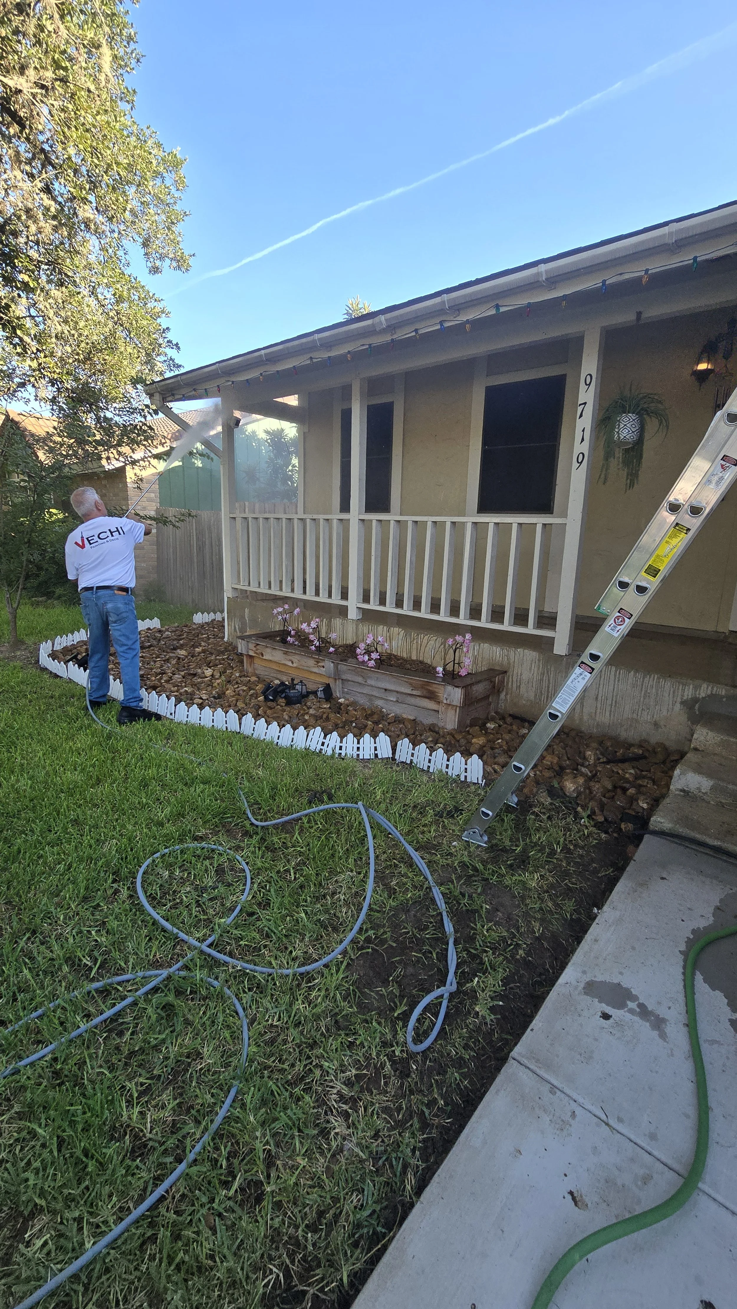 A person watering a garden bed in front of a house with a front porch, ladder leaning against the house, and a clear blue sky.
