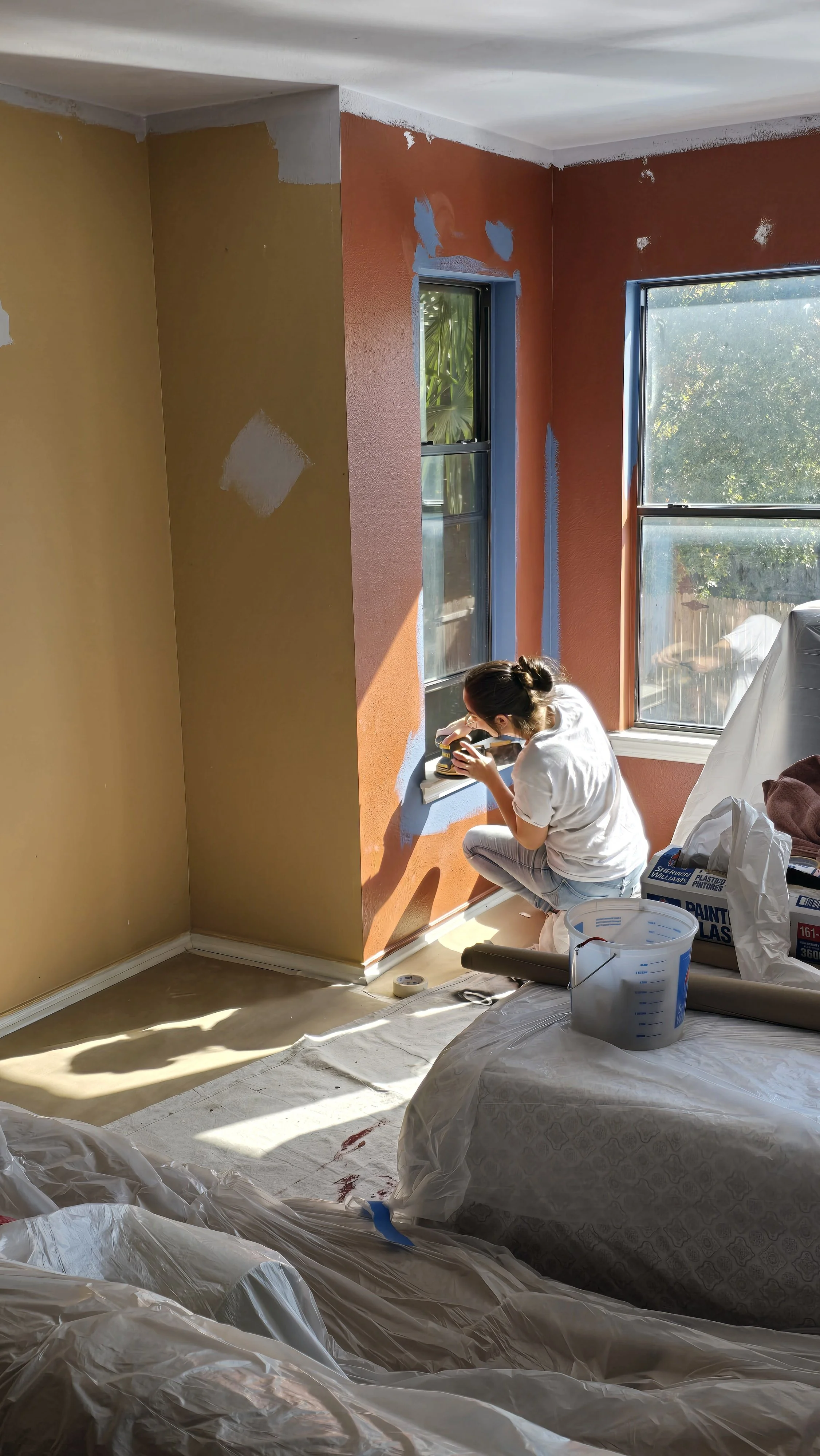 A woman working on painting a wall in a room with two windows, using painter's tape and a small roller, with painting supplies and plastic sheets covering furniture and the floor.