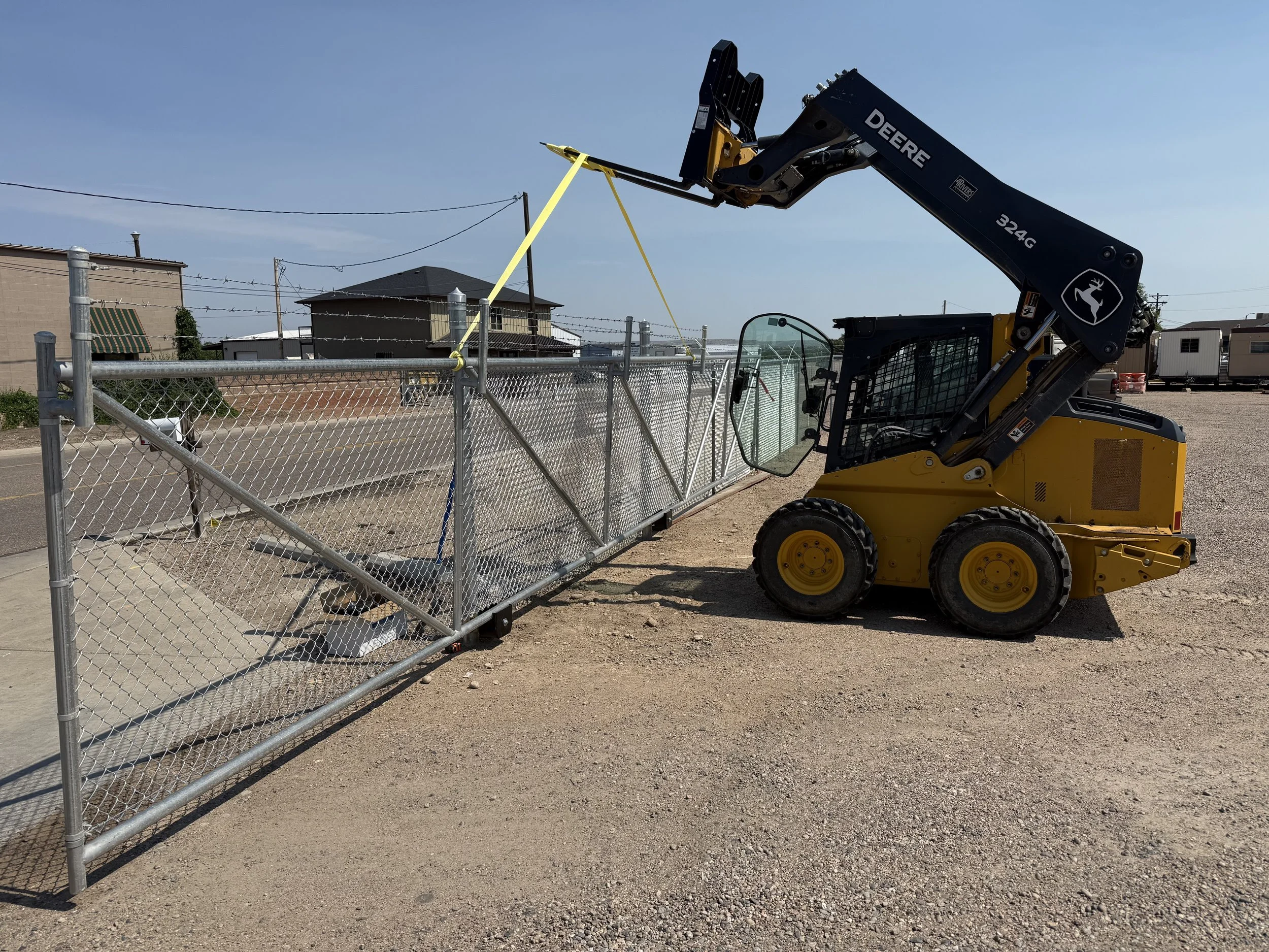 A yellow John Deere skid steer loader lifting a section of chain-link fence with yellow straps in an outdoor area on a sunny day.