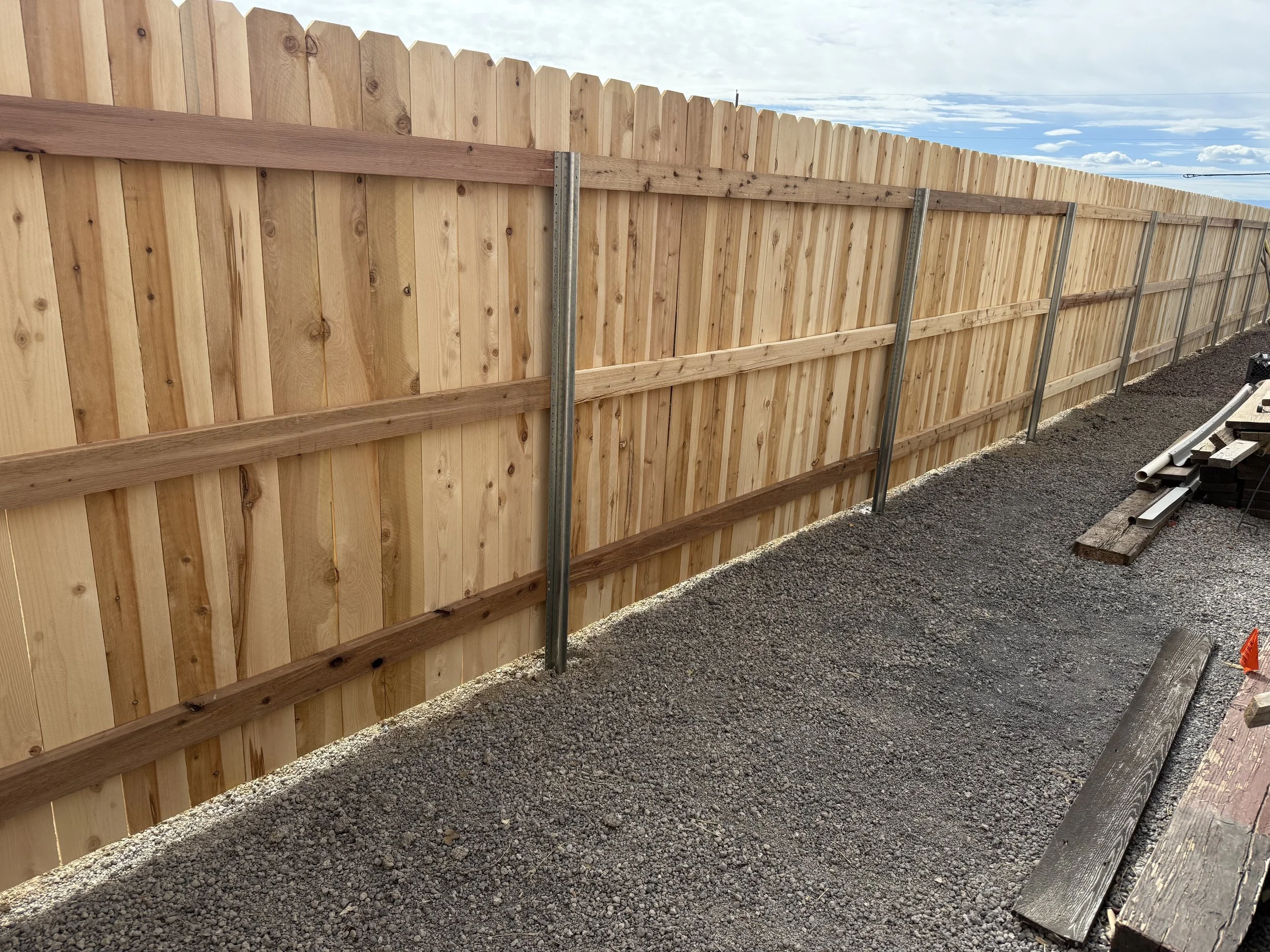 New wooden privacy fence under construction with metal posts, beside a gravel pathway, with construction materials on the side.