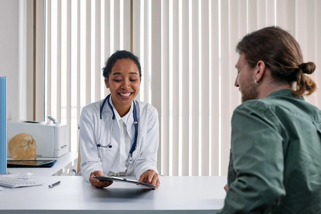 A doctor leans forward attentively while speaking with a patient during a medical consultation in a clinical office setting.