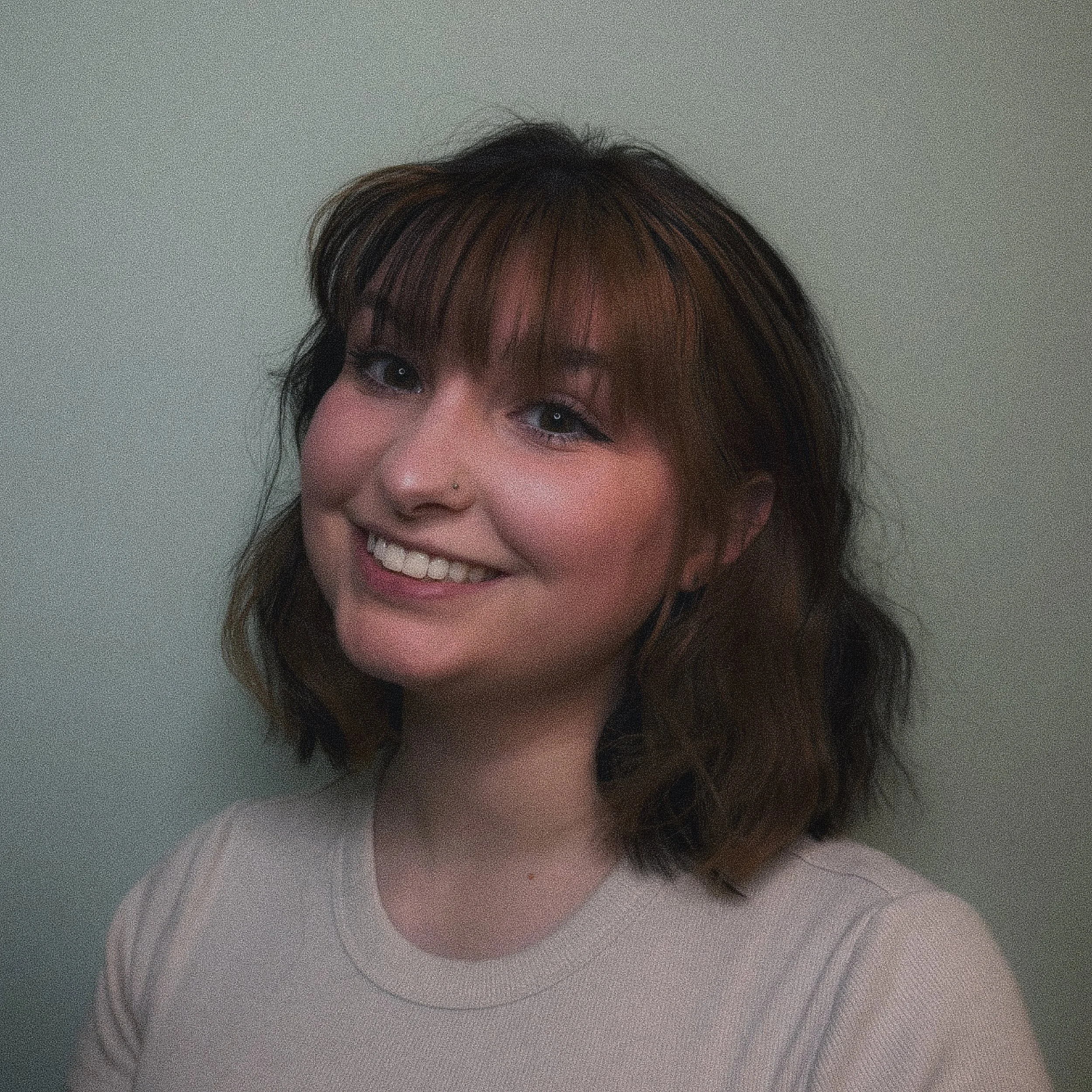 A young woman smiling with shoulder-length wavy brown hair, wearing a light-colored top, standing against a plain wall.