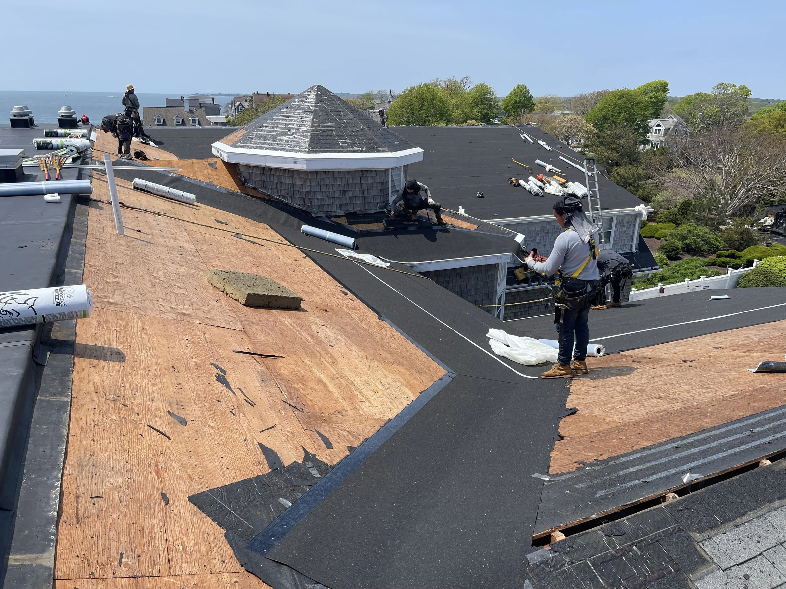 Roofers working on a residential roof, installing black roofing material, with some workers on the roof and one on the edge, surrounded by tools, shingles, and roofing materials, with a view of trees and houses in the background.