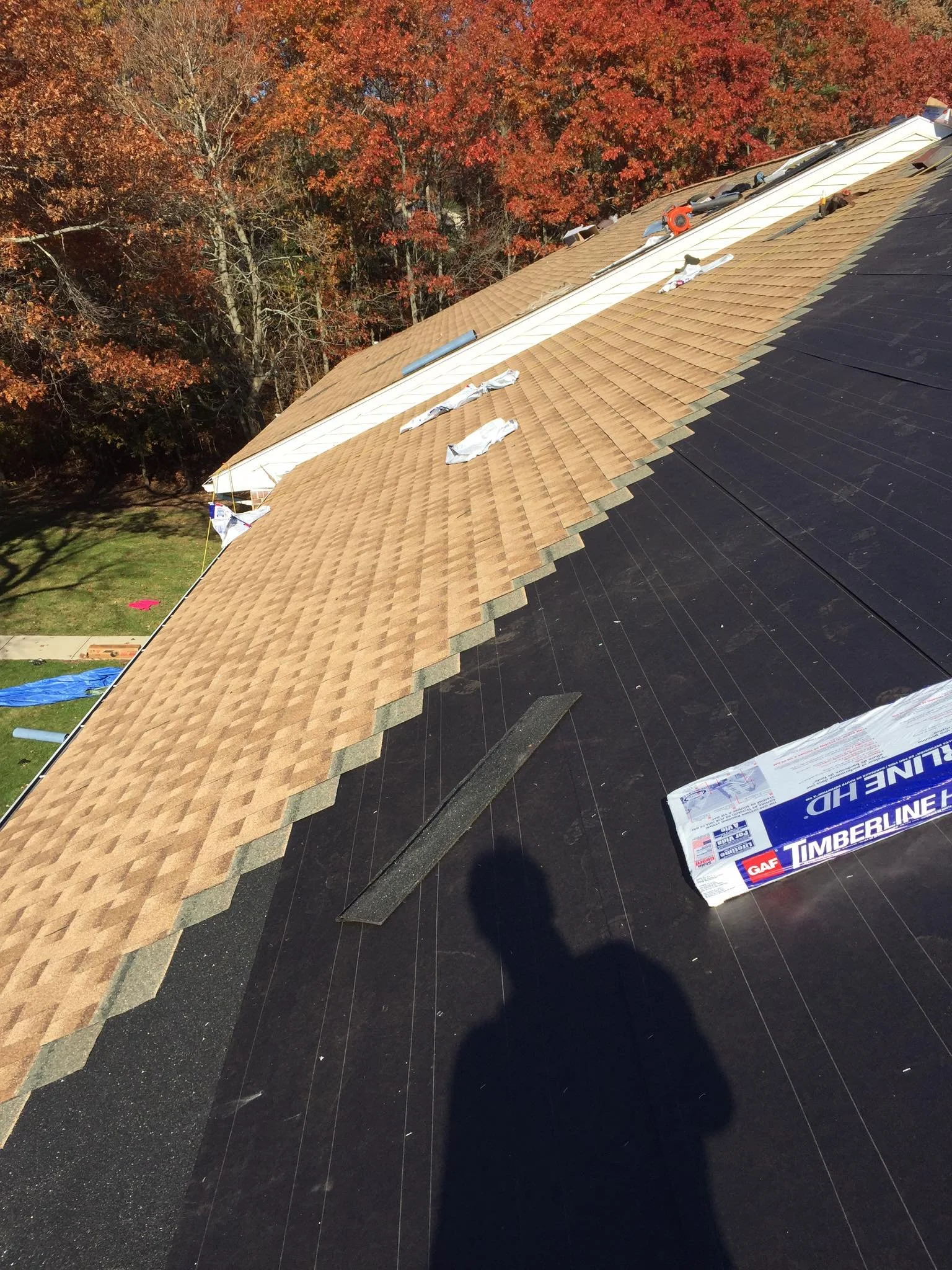 Roof with partially installed asphalt shingles, showing black underlayment, roofing tools, and materials, with trees with fall foliage in the background.
