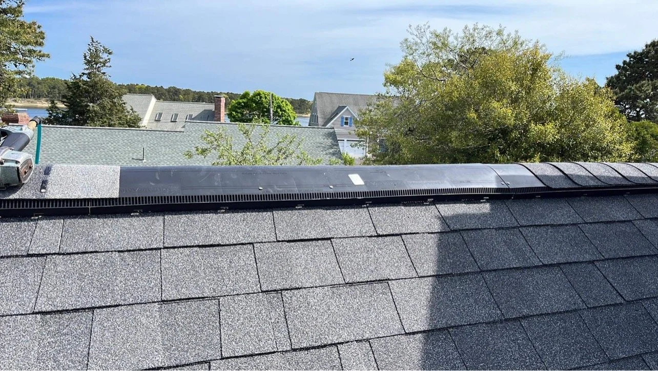 View of a house roof with shingles and a snow guard installed, with houses, trees, and a body of water in the background.