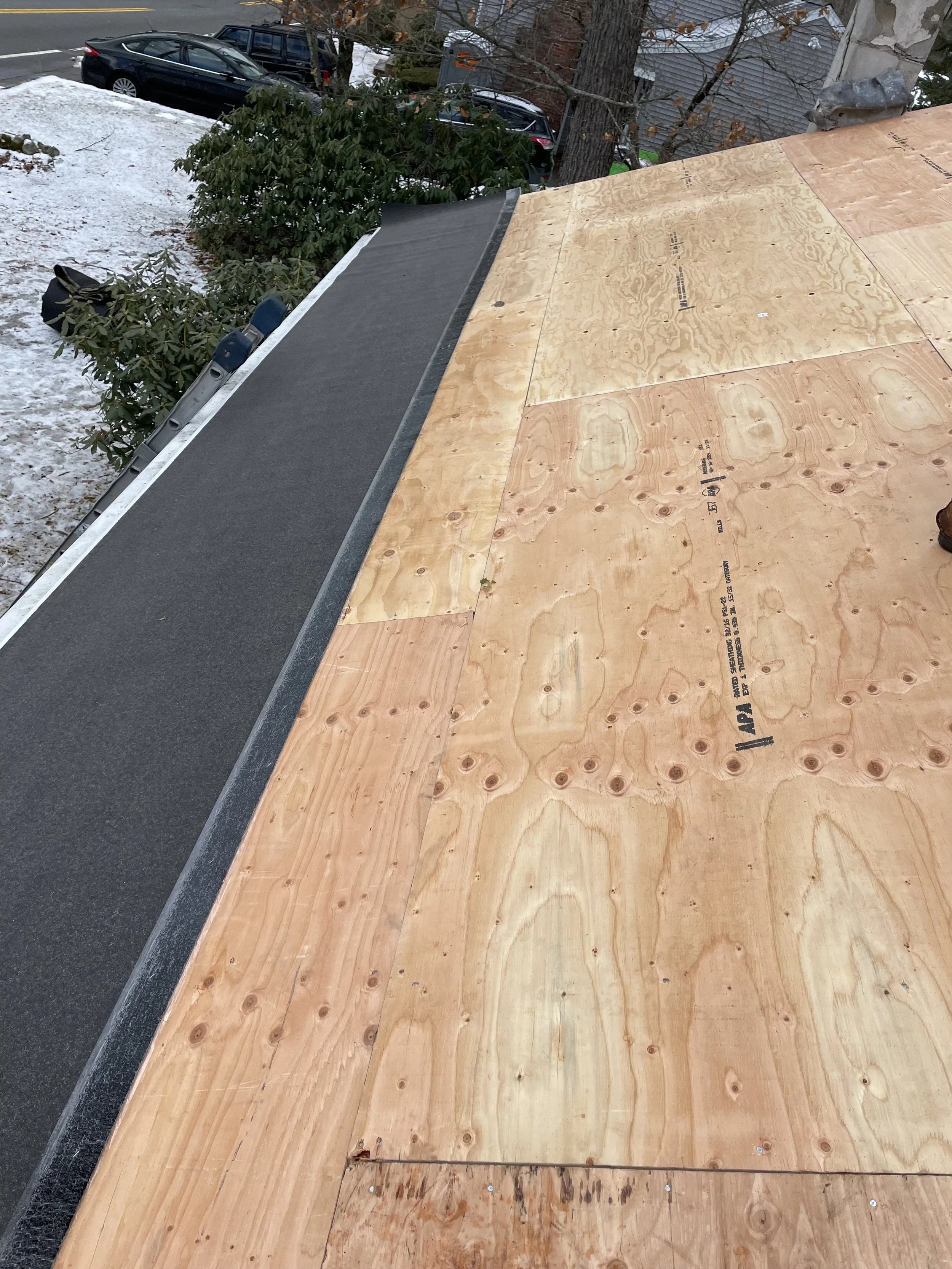 Wooden roof under construction with plywood sheets, next to a black roof shingle section, with cars parked and snow on the ground in the background.