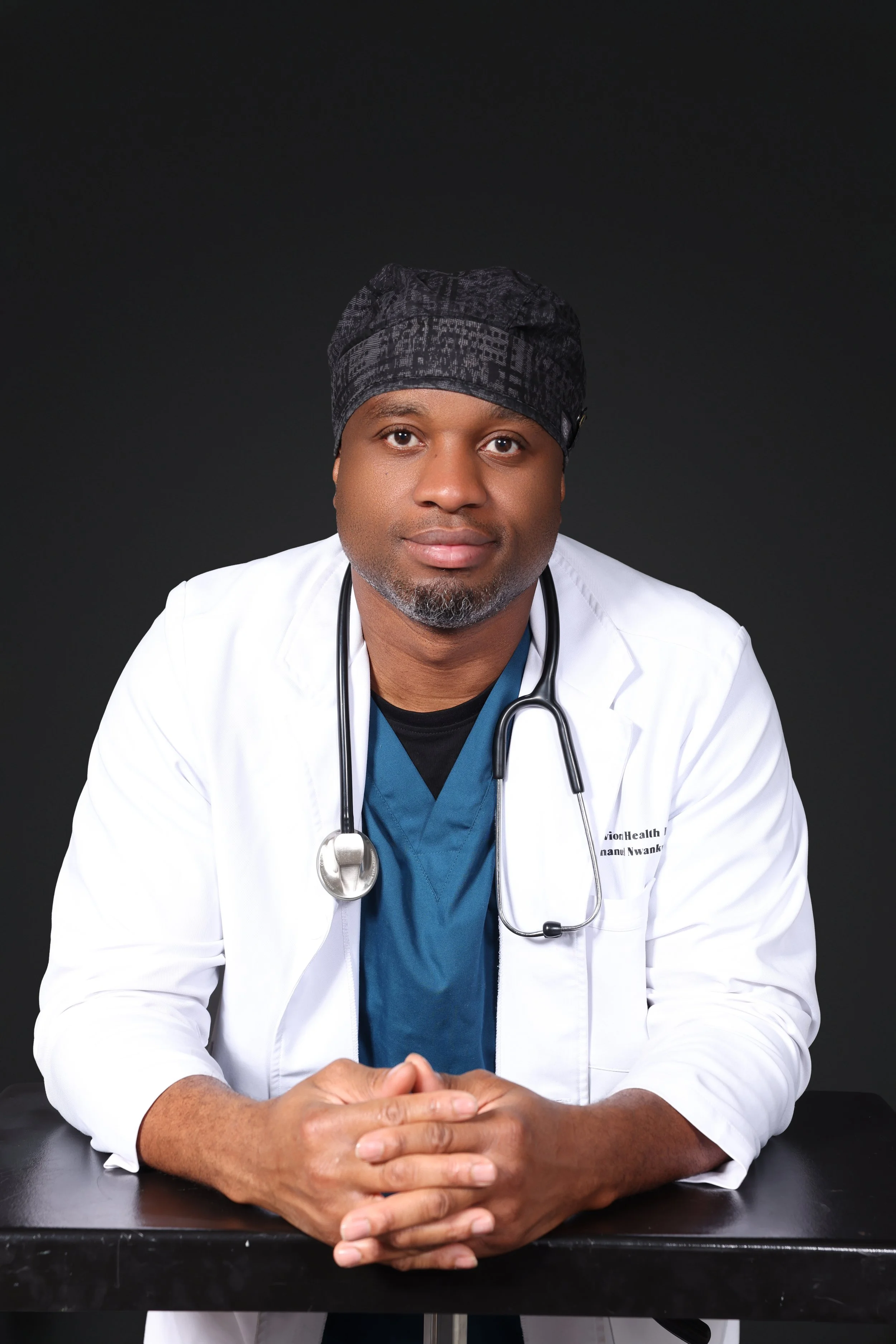 A male doctor in a white coat and blue scrubs with a stethoscope around his neck, sitting at a black table against a dark background.