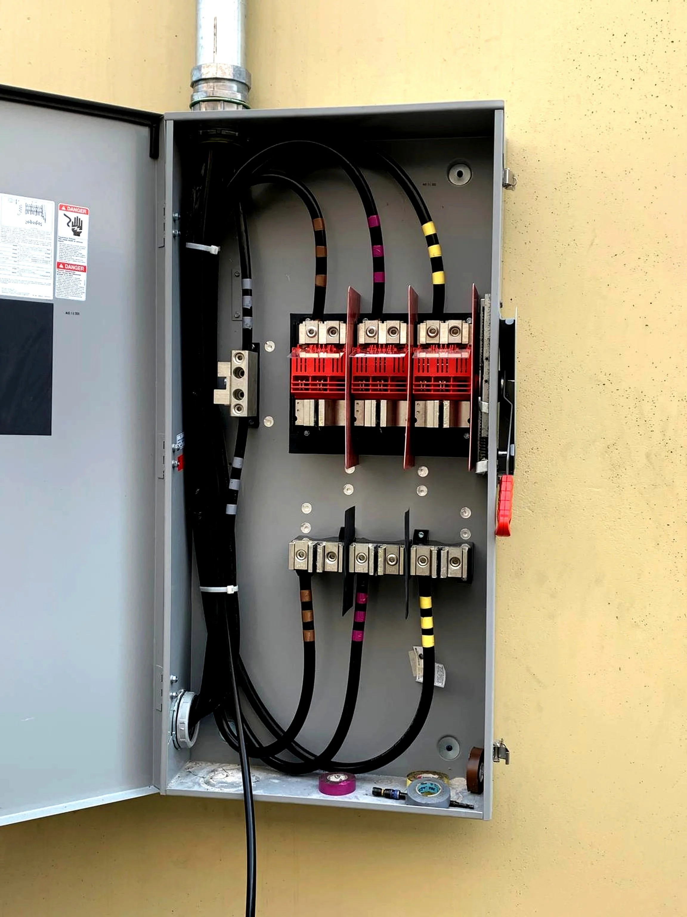 Electrical breaker panel with multiple colored wires and circuit breakers inside a metal box mounted on a yellow wall.
