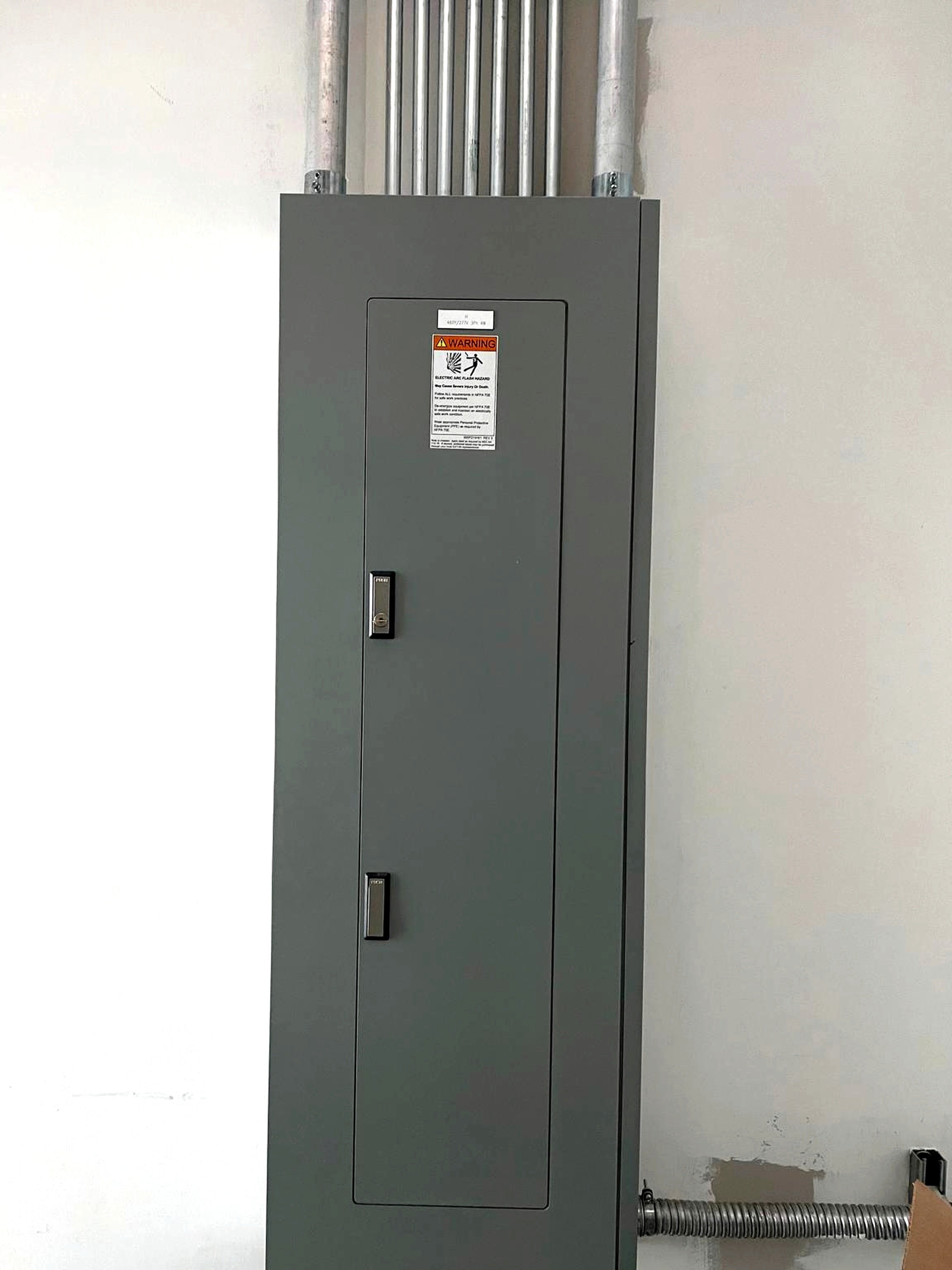 Electrical panel with warning label on a light-colored wall, with conduit pipes above and a flexible conduit on the floor.