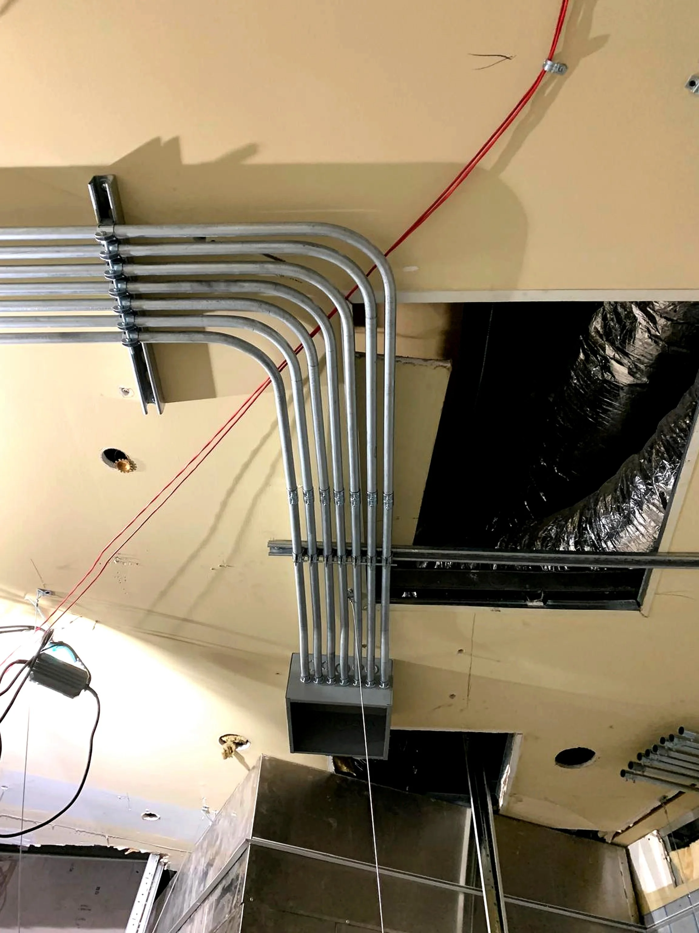 Metal electrical conduit pipes running from the ceiling into a rectangular box, with some red wires and black ductwork visible in an unfinished building space.
