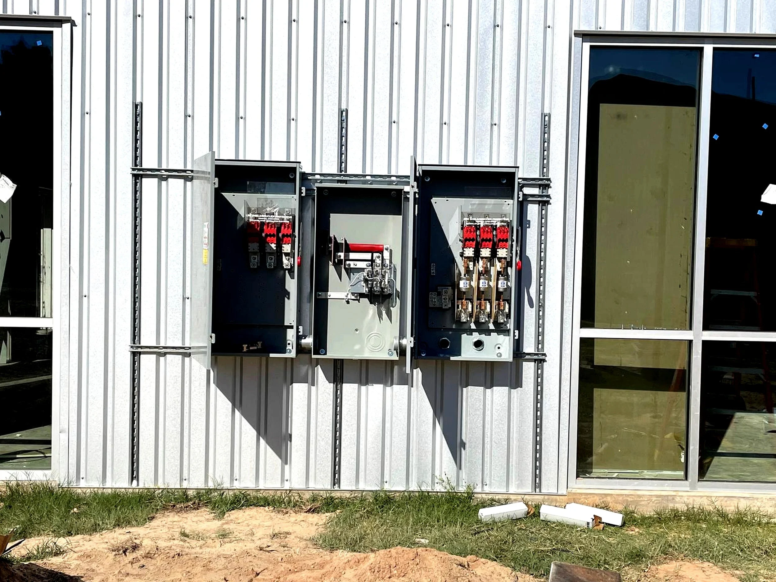 Three electrical meter boxes on a side of a building with metal siding, one of which is open showing electrical components inside.