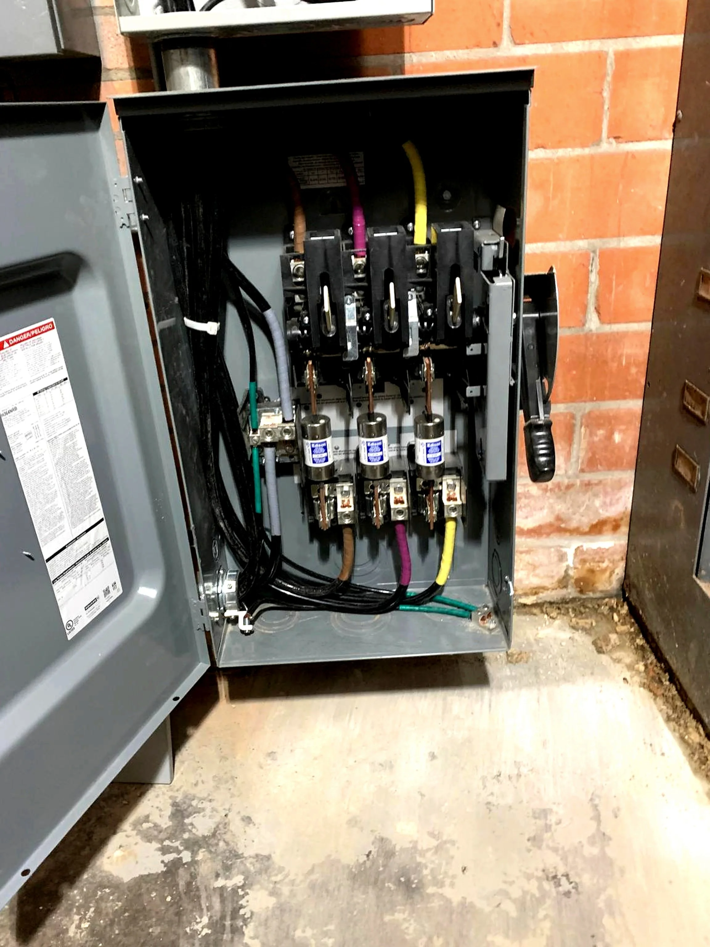 Open electrical panel with visible wiring, circuit breakers, and fuses mounted inside, located in a basement or utility area with brick wall and concrete floor.