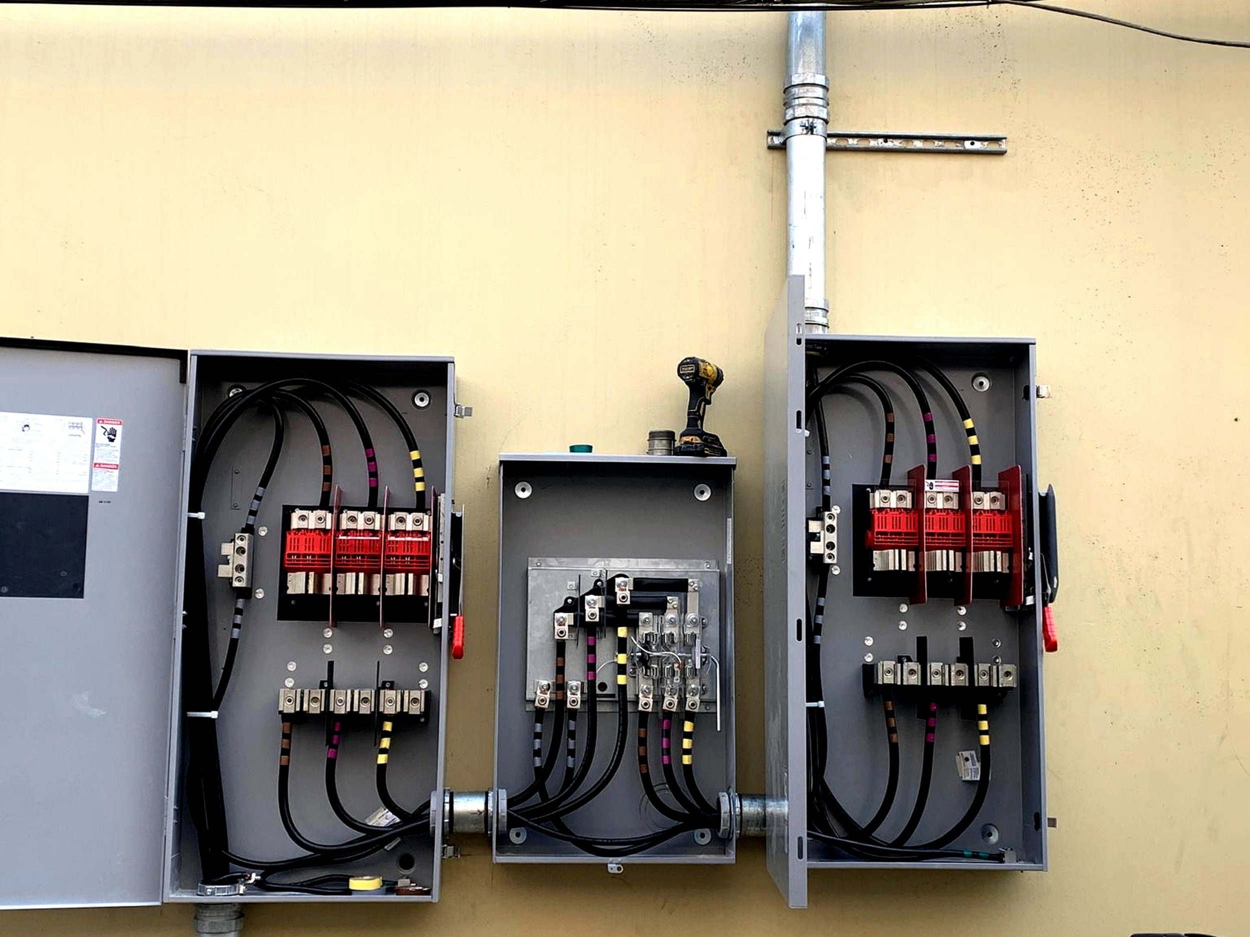 Open electrical panels with black and yellow cables on a yellow wall, with a cordless drill on top of a small open box