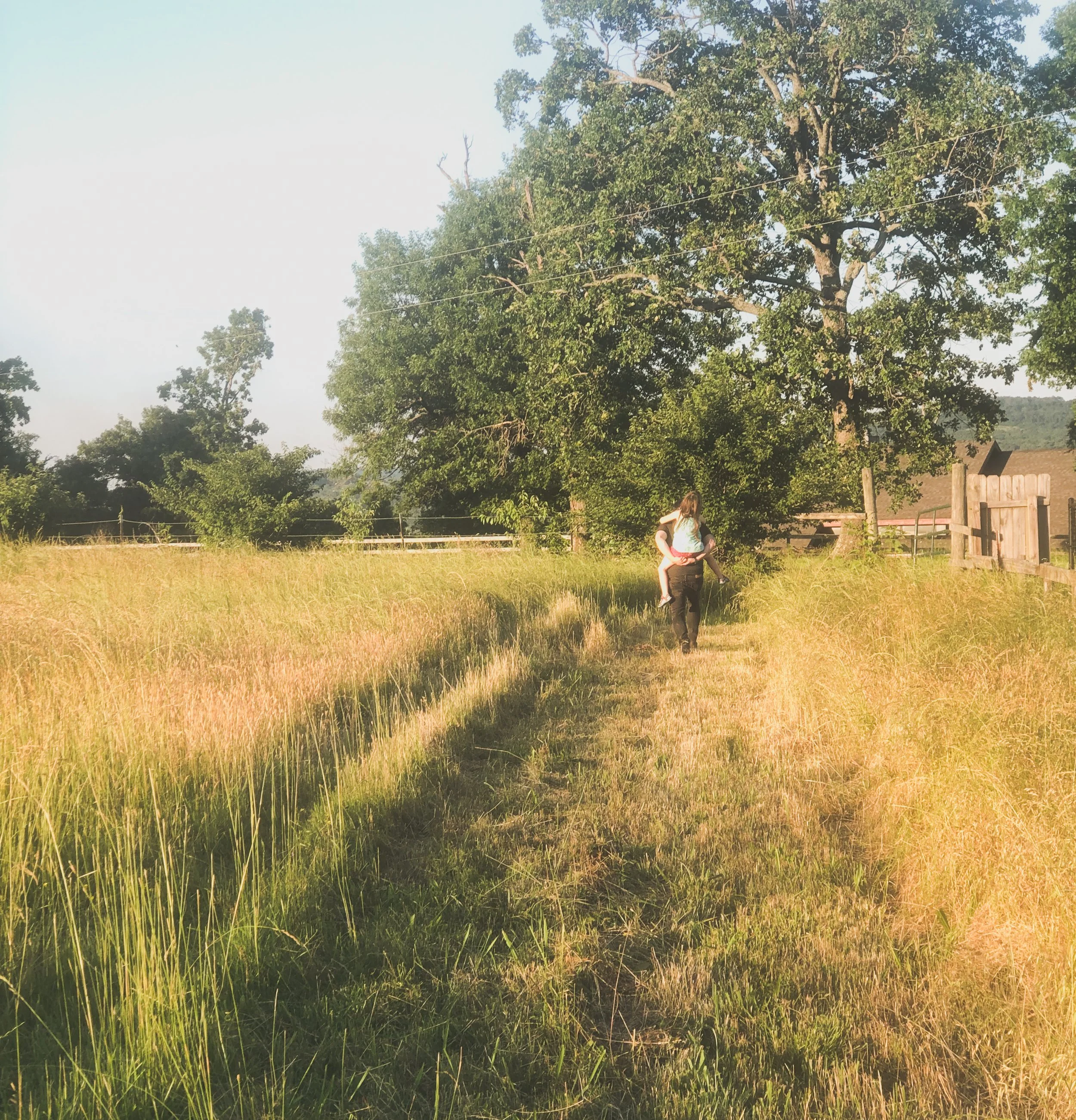 A person carrying a young girl on their shoulders walking along a grassy trail in a field with tall grass and trees, with a wooden fence and house in the background, during late afternoon sunlight.