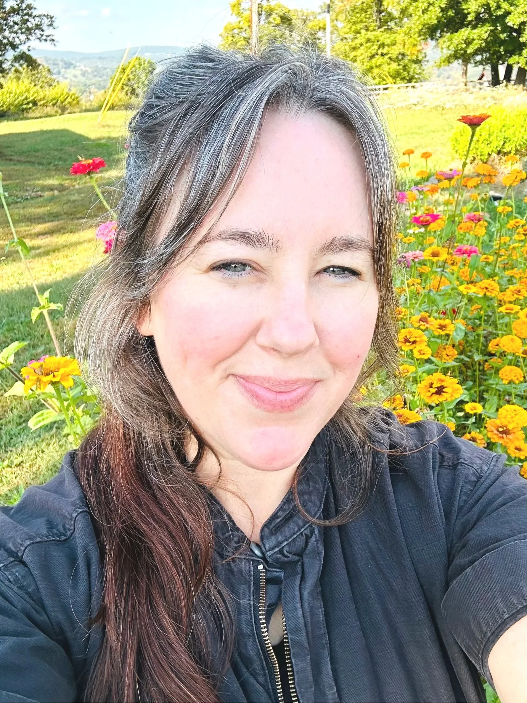 A woman taking a selfie outdoors in a garden filled with colorful flowers, woodland behind, and distant mountains in the background on a sunny day.