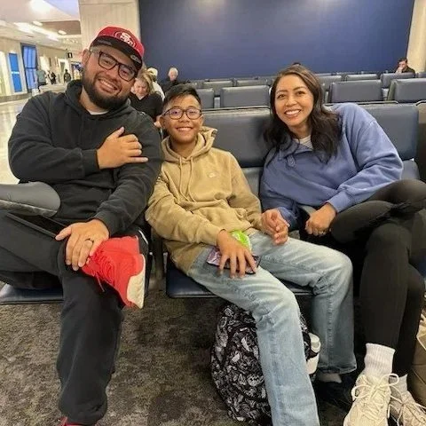 Three people sitting in an airport waiting area, smiling and posing for the photo. The group includes a man, a woman, and a young boy.