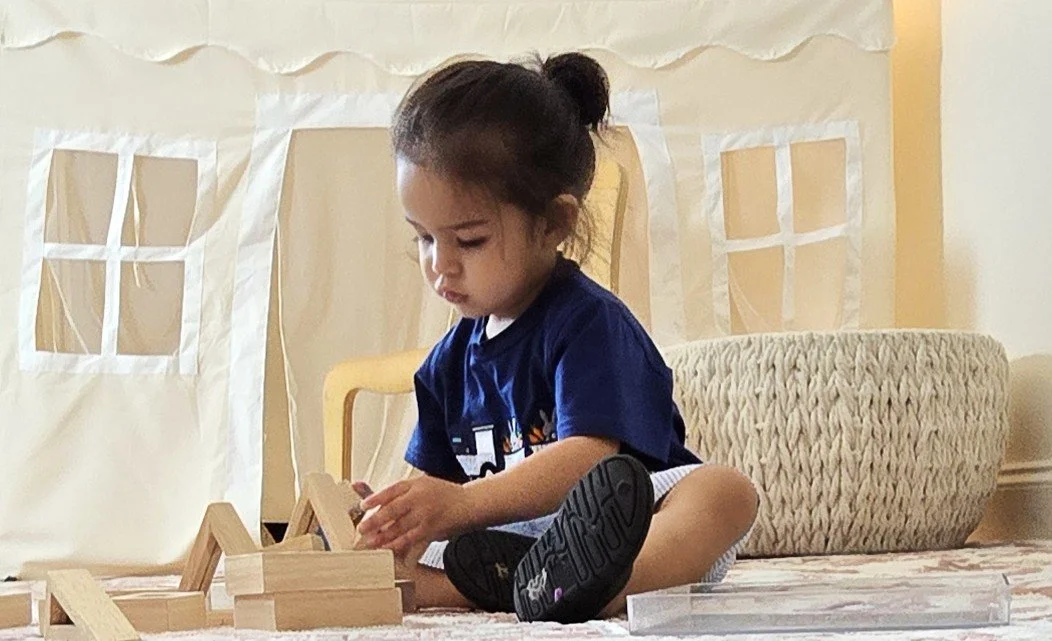A young boy sitting on the floor playing with wooden blocks inside a room with a cream-colored curtain and large woven ottoman behind her.