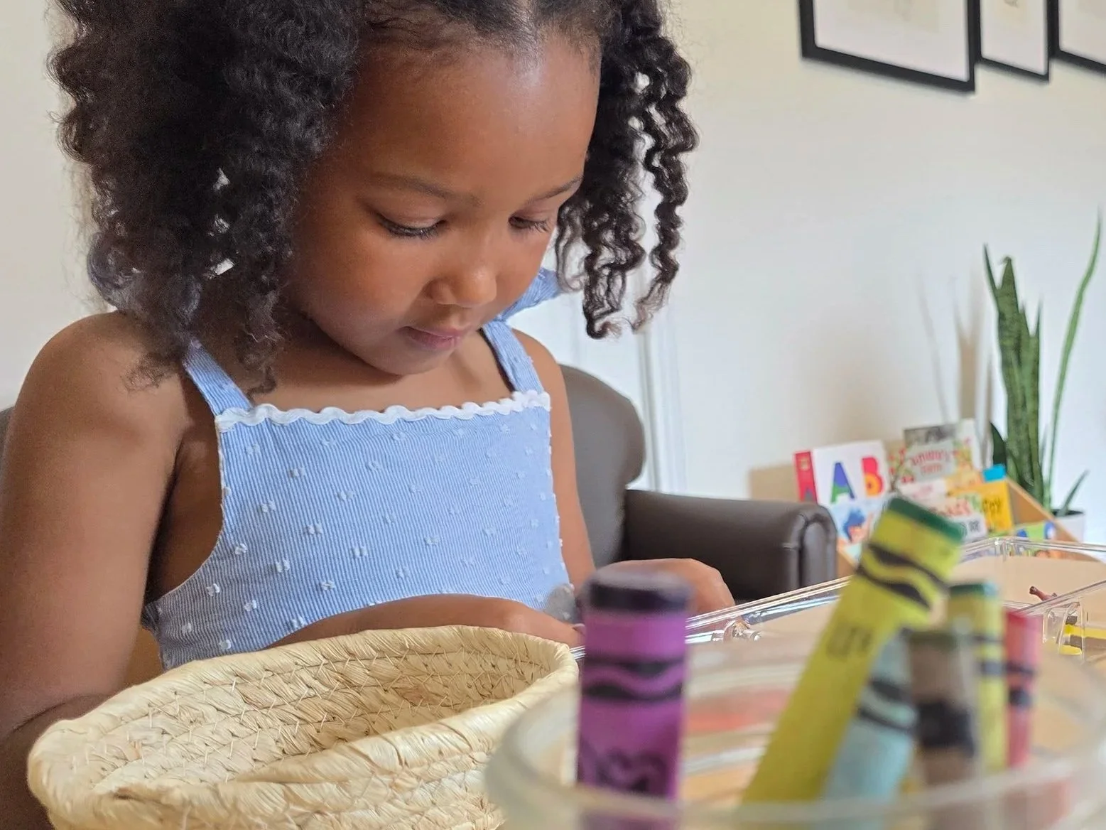 A young girl with curly hair looking down at a table with art supplies, in a room with books and houseplants.