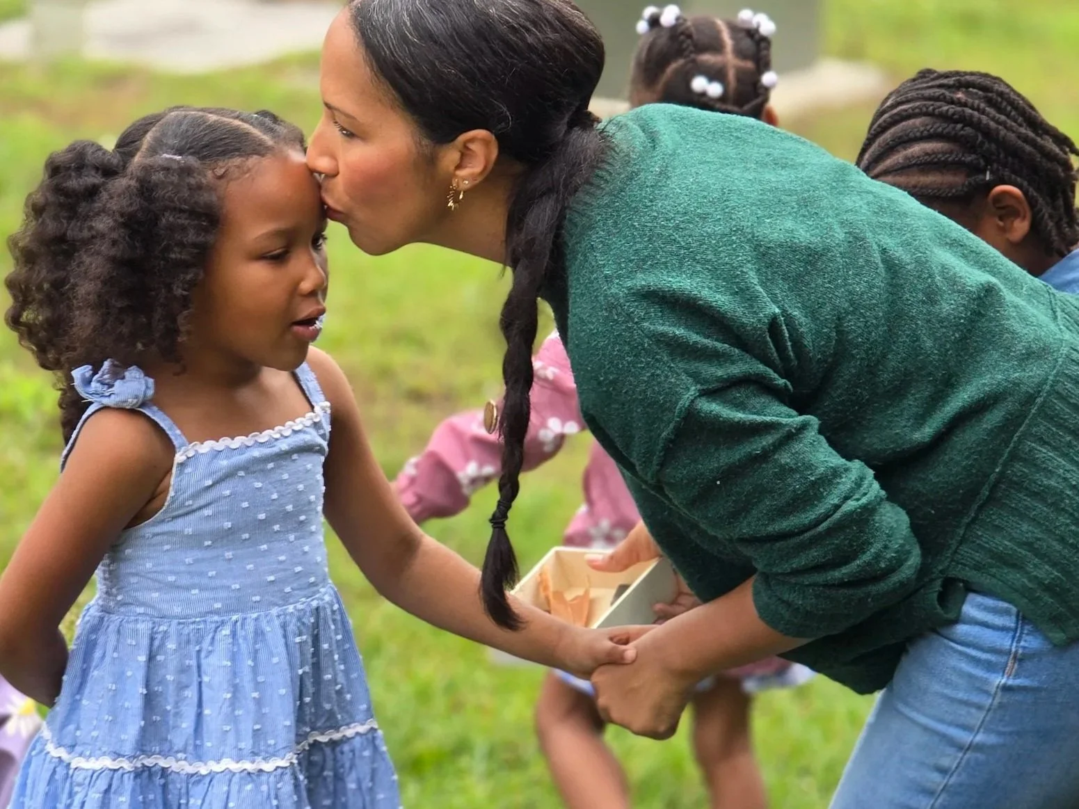 A woman with dark hair and a green sweater is kissing a young girl with curly hair and a light blue dress on the forehead in an outdoor setting, with other children in the background.