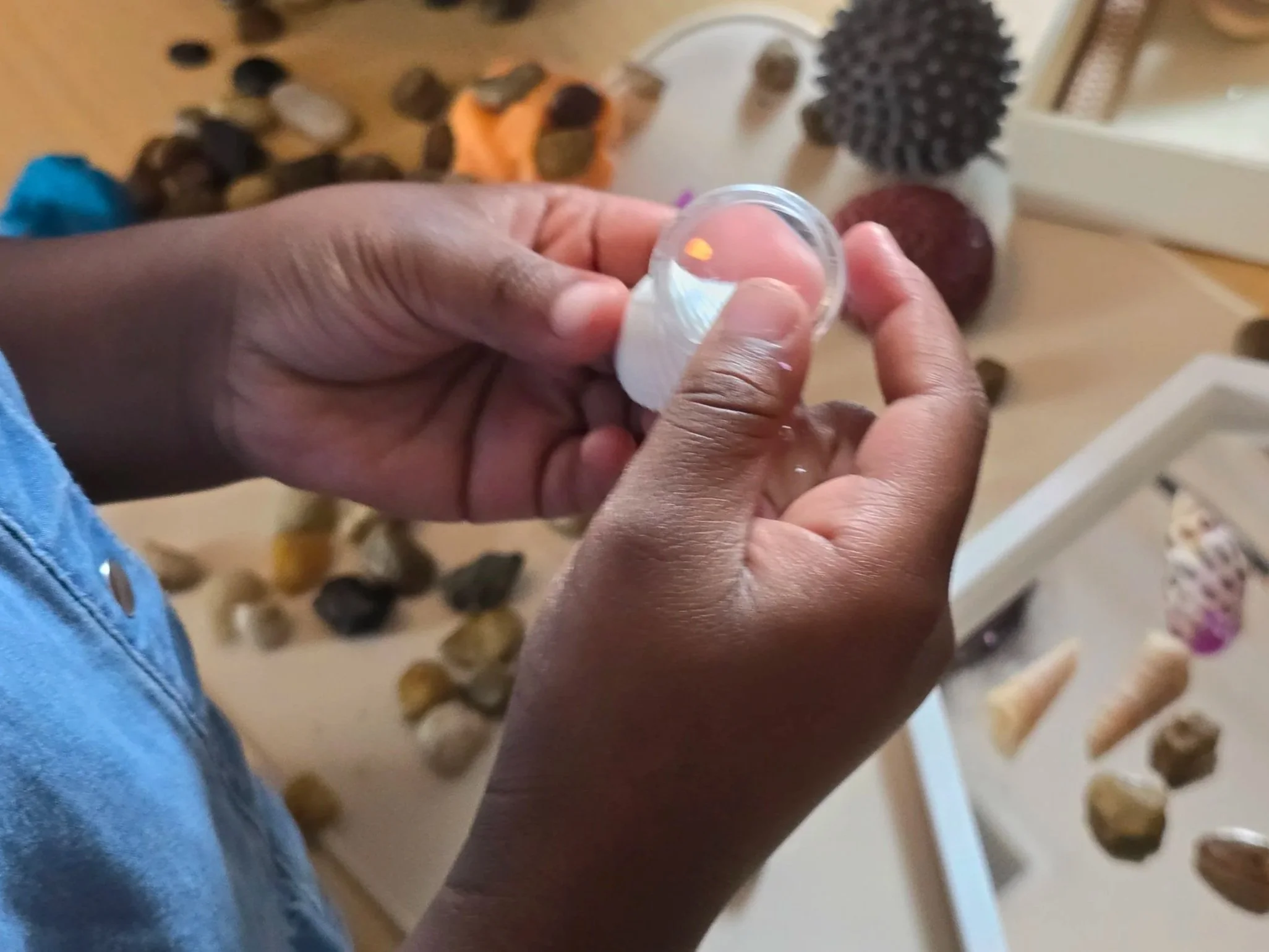 Close-up of a person's hands holding a small transparent plastic container with a white lid, with a colorful background of objects including rocks, a black textured item, and a red object.