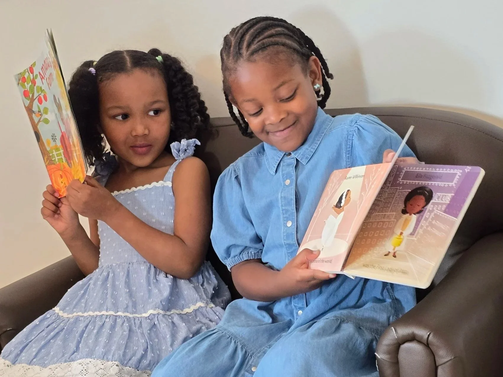 Two young girls sitting together on a brown armchair, reading books. The girl on the left has curly hair in pigtails wearing a light blue dress with white polka dots and ruffled sleeves. The girl on the right has braided hair with beads, wearing a bl