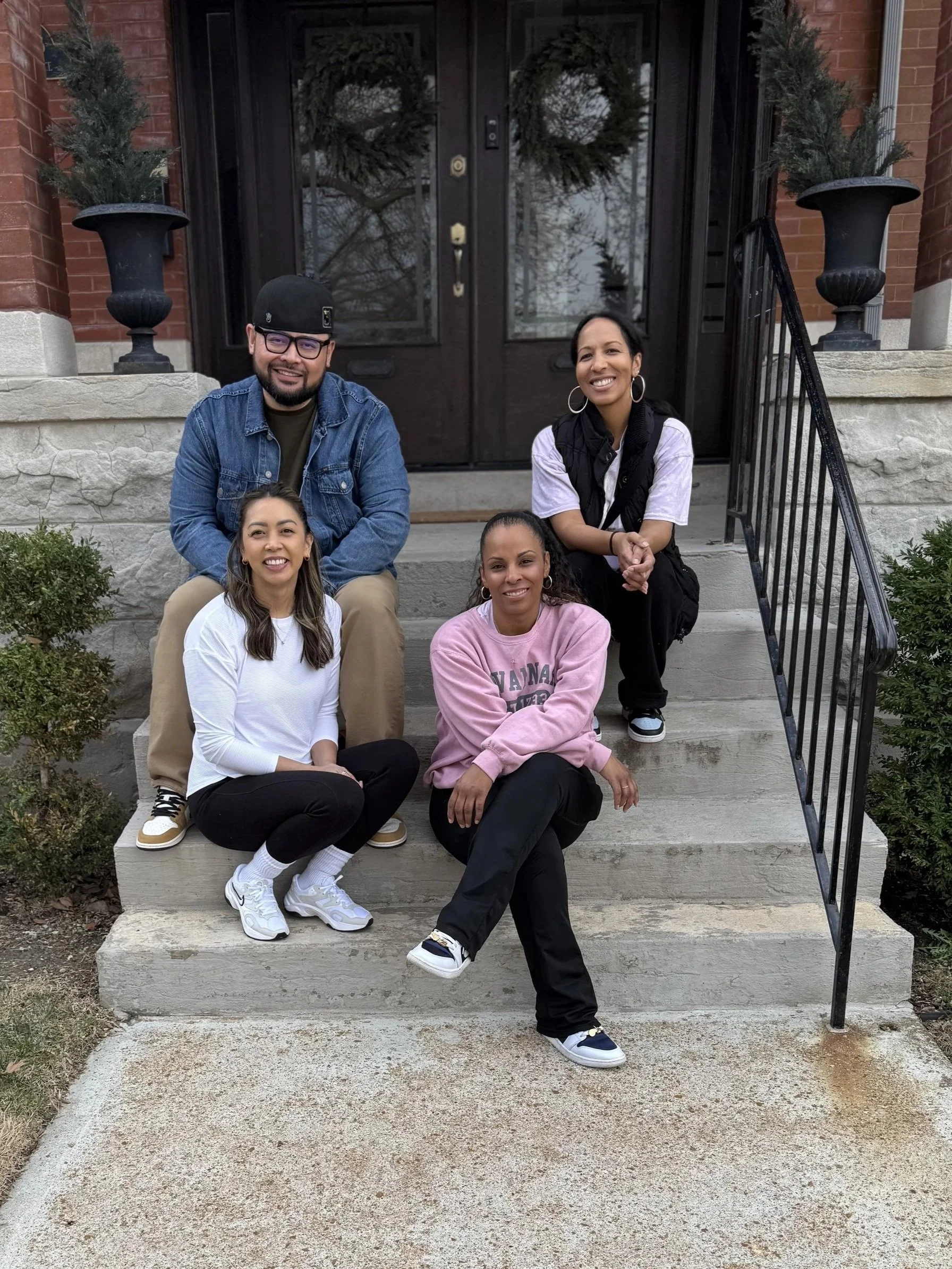 Group of five diverse friends sitting on the front steps of a brick house with glass-paneled double doors decorated with Christmas wreaths and two large potted plants on either side, smiling and enjoying each other's company.