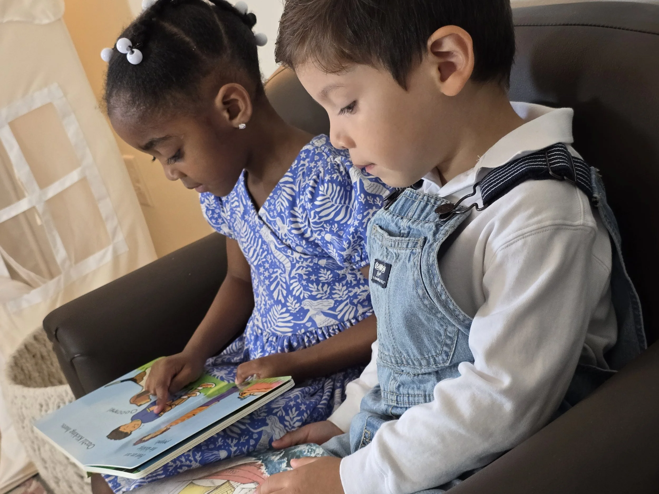 Two children, a girl and a boy, sitting close together on a couch and looking at a colorful children's book.