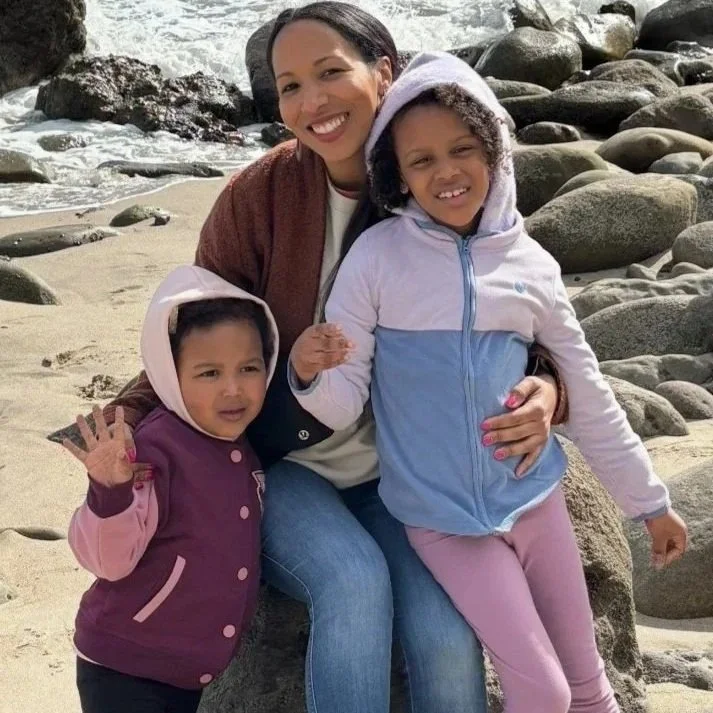 A woman with two young girls on a beach with rocks and sand, smiling. The women and girls are dressed in hooded jackets.