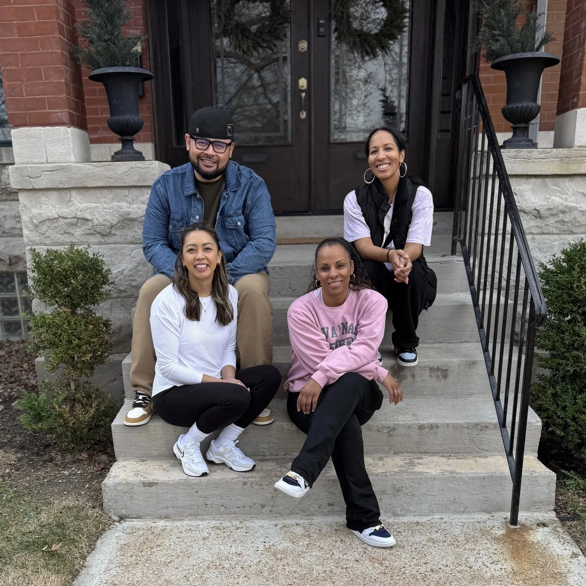 Group of five diverse people sitting on front steps of a house, smiling at camera, with brick house and trees in background.